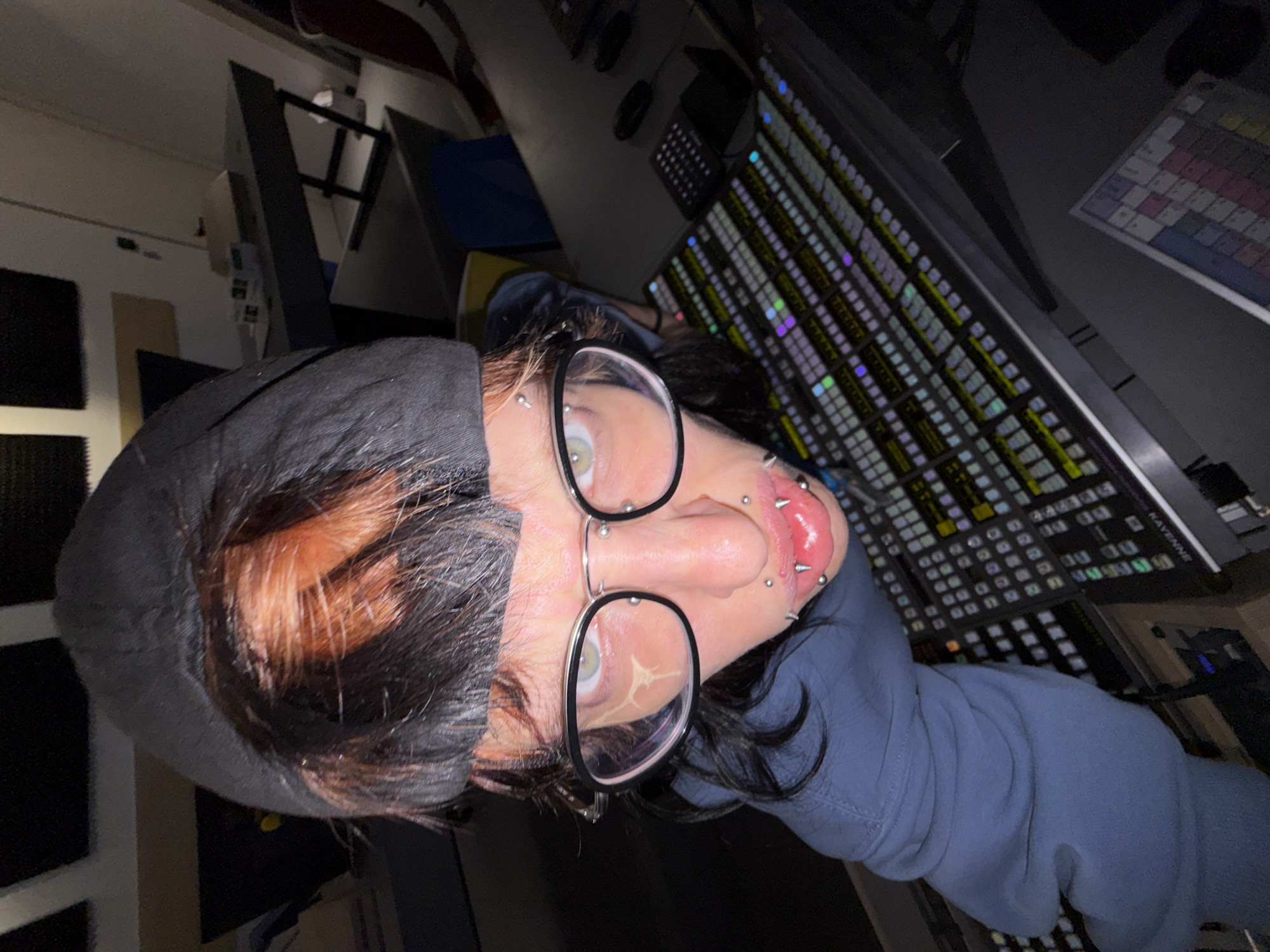 A person wearing glasses and multiple piercings in and around their left ear, eyebrow, and lower lip is angled slightly downward toward a large electronic control panel. The person is wearing a dark-colored hooded garment. The control panel, bearing the name "KAYENNE," consists of numerous illuminated buttons and switches arranged in a grid pattern. A computer keyboard lies to the left of the control panel, and a periodic table poster is visible in the upper right corner. The background includes additional equipment and structural elements of a room, indicating an indoor setting. The location of the scene is not discernible as Sofia, Bulgaria.