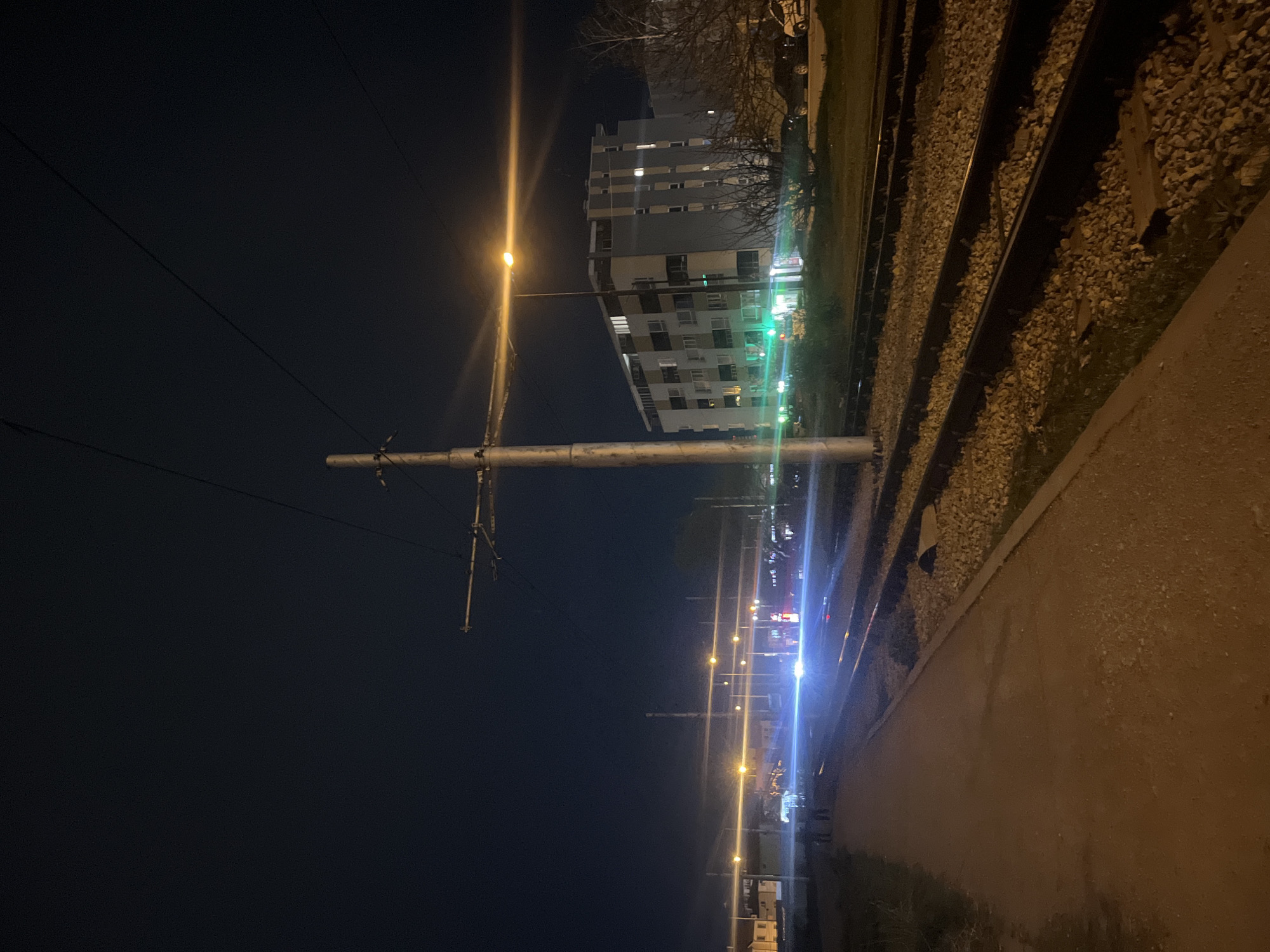 The image depicts a nighttime scene in Sofia, Bulgaria, near a tram line. A tall, narrow apartment building with multiple floors is visible in the background. A series of streetlights stretches into the distance. Tram tracks run alongside a concrete pathway. A tram pole is situated in the foreground, with overhead wires extending across the dark sky.