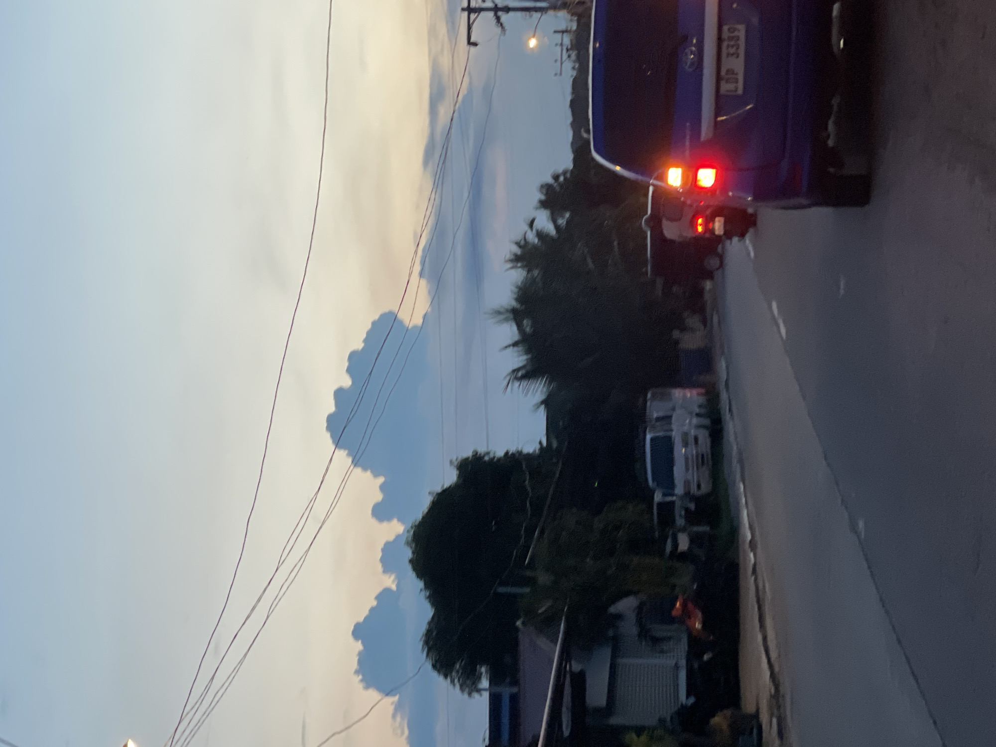 The image depicts a street scene at dusk in Davao City, Philippines, likely along a suburban or residential road. The sky is a gradient of pale blue and light orange, with distinct, fluffy clouds illuminated by the setting sun. Power lines are visible crisscrossing the sky.
On the left side of the road, there are buildings with a reddish-brown roof and some white structures. Trees and foliage, including what appears to be palm trees, line the area behind the buildings.
The road itself is asphalt and appears to be a two-lane road, with vehicles traveling in the same direction. In the foreground, a blue SUV is prominently featured, its brake lights illuminated. The license plate of the SUV is visible and reads "LBP 3389". Behind the SUV, a motorcycle with its red taillight on is visible, followed by other vehicles, including a white van. The overall lighting suggests it is either early morning or late evening.