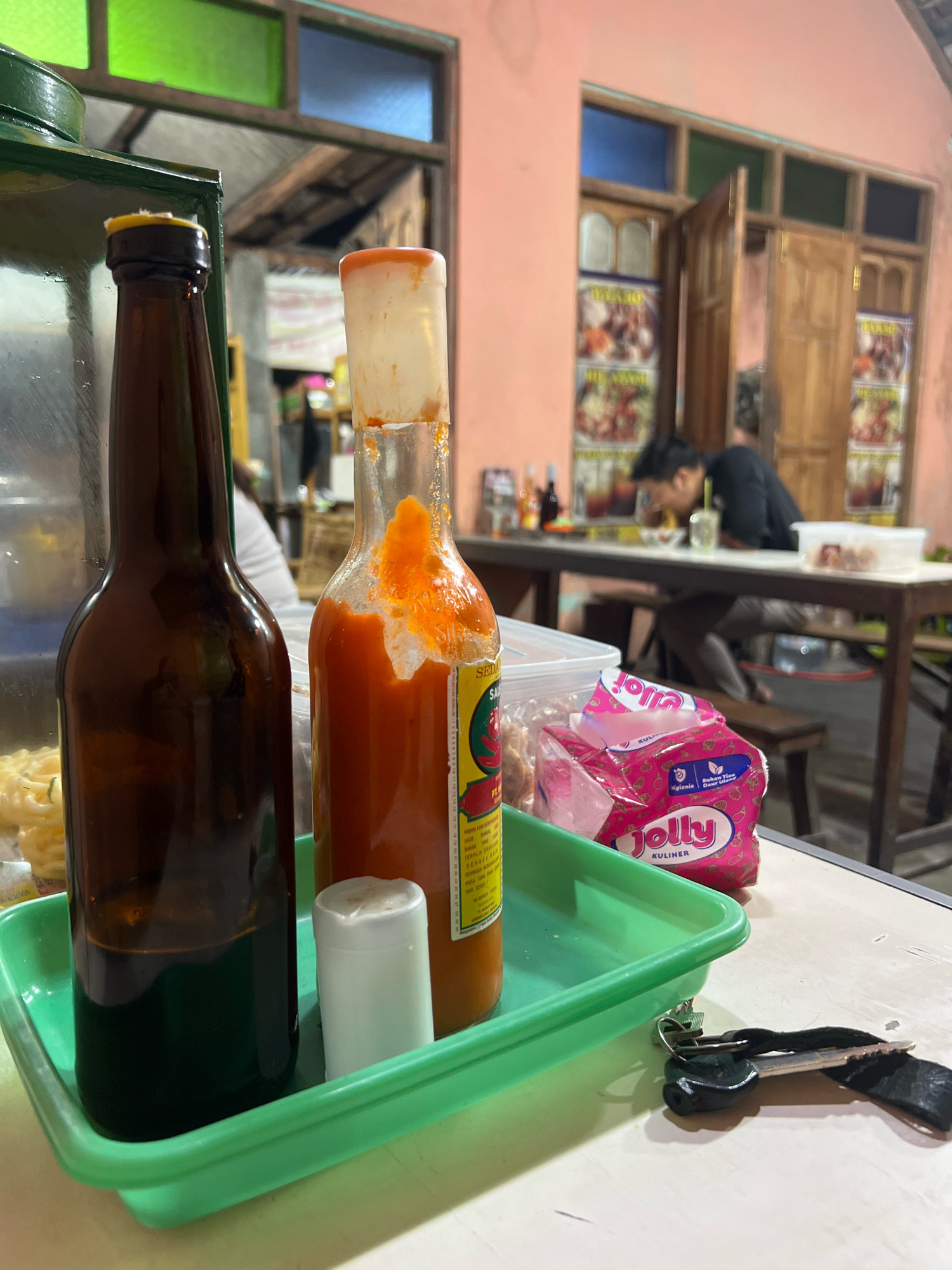 In Sleman, Indonesia, a close-up shot captures a table setting at what appears to be a casual eatery or food stall. Dominating the foreground are two bottles, one a dark amber glass bottle with a dark liquid, and the other a clear glass bottle containing bright orange chili sauce, its cap and neck stained with residue. A small white cap or dispenser rests beside them, all contained within a bright green plastic tray. To the right of the tray, a packet of pink tissues labeled "Jolly KULINER" is visible. In the background, a person with dark hair is seated at a wooden table, hunched over a bowl, seemingly engaged in eating. The environment suggests an outdoor or semi-outdoor setting, with a building featuring windows with colored panes and wooden doors in the background. A person is seated at a table, eating. The overall scene portrays a moment of casual dining in a local Indonesian establishment.