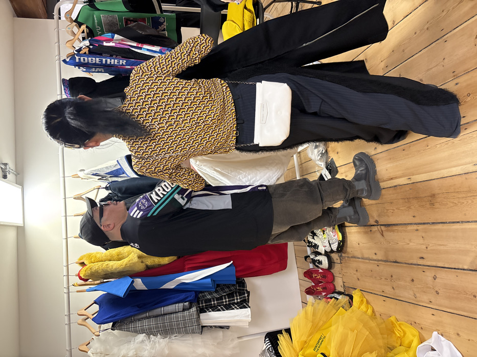 The scene depicts an indoor area resembling a fashion showroom or backstage space in the city of London, United Kingdom. Two individuals are present, standing on a light-colored wooden floor beside a white metal clothing rack that extends horizontally across the frame. The walls are white, and a rectangular fluorescent light fixture is visible on the ceiling.
On the left, a male individual is seen from his left profile, wearing a black baseball cap, dark sunglasses, a black long-sleeved shirt with purple, teal, and white accents, and the partial text "KROL" on the sleeve. He wears dark grey baggy pants and dark grey chunky-soled boots. He faces towards the clothing rack.
On the right, a female individual is viewed from the back. She wears a long-sleeved top with a yellow and dark geometric pattern, dark pinstriped wide-leg trousers featuring prominent black furry panels along the outer seams, and a white rectangular shoulder bag with a silver chain strap. Her dark hair is pulled back into a ponytail. She is holding a long, dark garment, possibly a coat or dress, in her hands, appearing to be in the process of examining or preparing it.
The clothing rack holds a diverse selection of garments, including a white ruffled item, a blue dress, a red dress, a yellow faux fur coat, plaid clothing, and graphic t-shirts, one of which displays the text "TOGETHER WE ARE". On the wooden floor beneath the rack and to the left, several pairs of shoes are visible, including red athletic shoes, white and black sneakers, and other colorful trainers. A bright yellow ruffled fabric item rests on the floor to the far left.