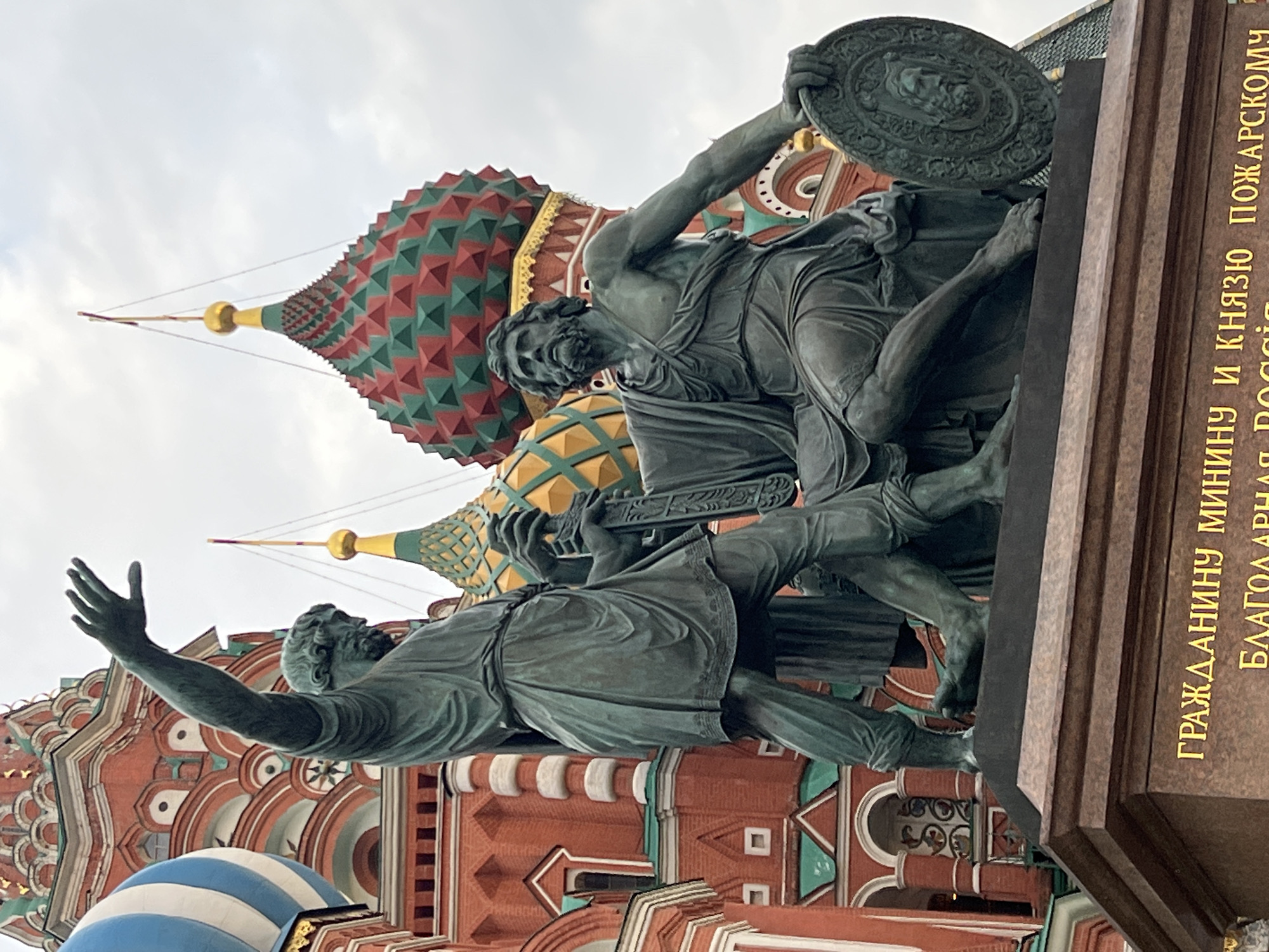 The image shows the Monument to Minin and Pozharsky in front of St. Basil's Cathedral in Moscow, Russia. The monument depicts Kuzma Minin gesturing towards Dmitry Pozharsky, who is holding a shield. The inscription on the monument's base is in Russian. The background features the colorful domes of St. Basil's Cathedral and a cloudy sky.