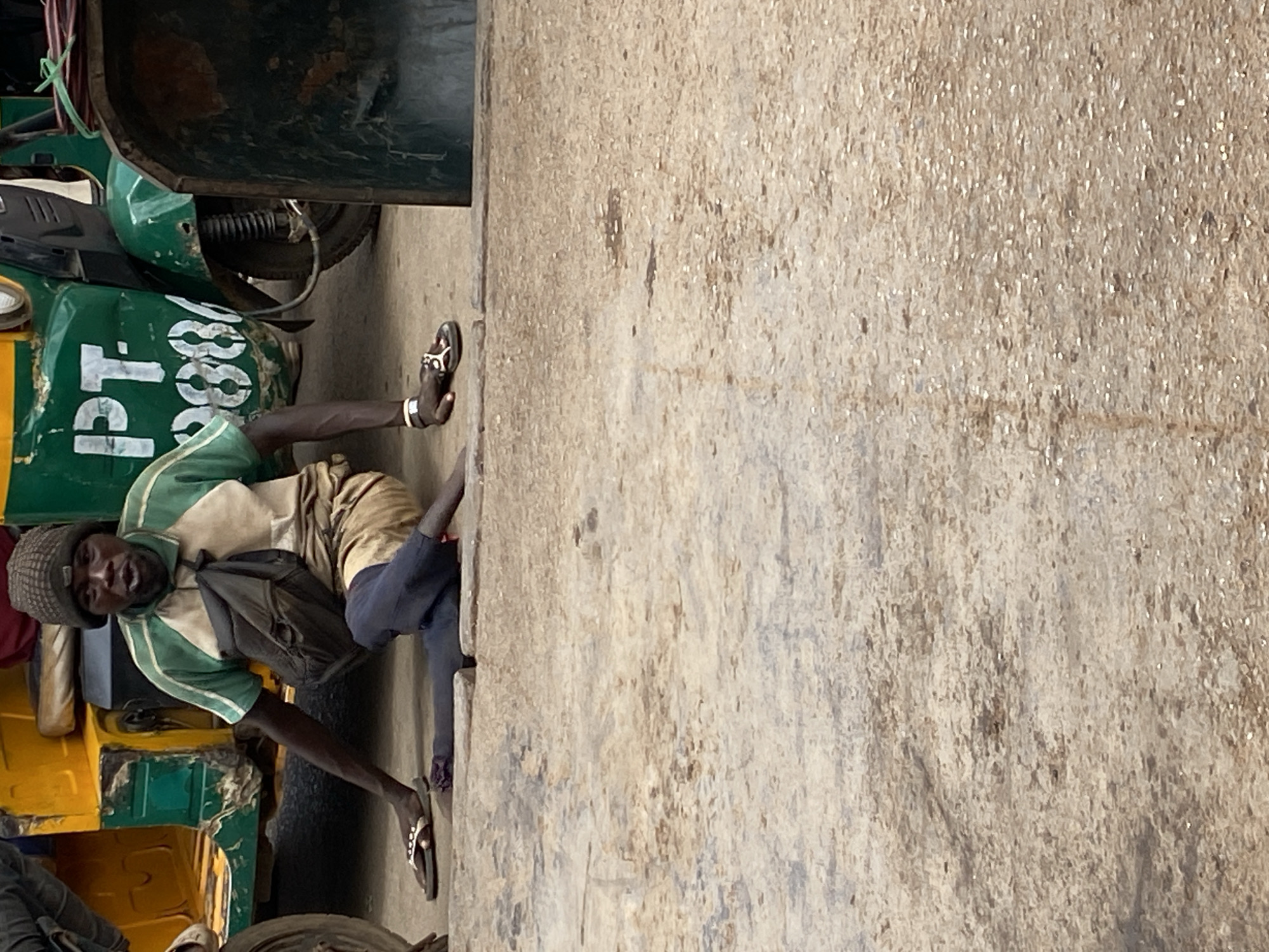 A man is positioned adjacent to a wall in Bukuru, Nigeria, next to an auto-rickshaw. The man is dark-skinned and wears a grey beanie, a shirt with green and white stripes, dark-blue pants, and sandals. He also carries a dark backpack. The auto-rickshaw is predominantly green and yellow, with the inscription "PT 8888" on its side. The wall is concrete, showing signs of wear and discoloration. The overall scene suggests a common urban environment.
