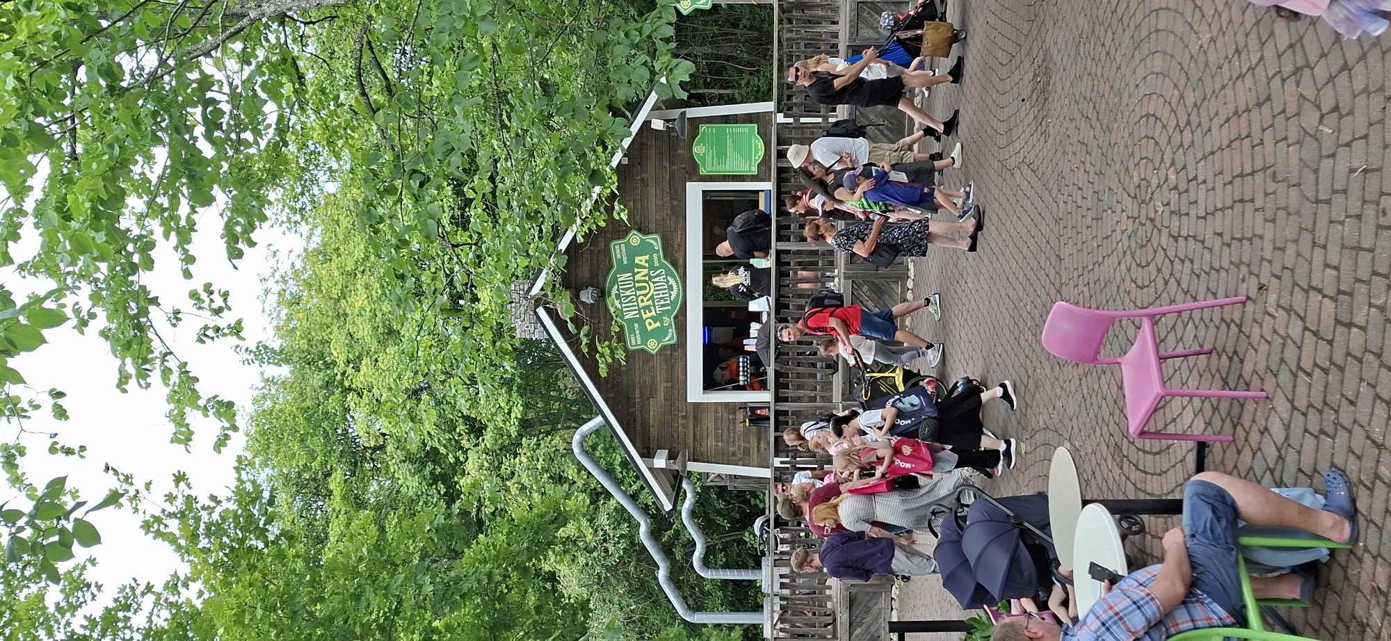 The image depicts an outdoor scene at a food establishment, likely in Naantali, Finland, given the Finnish text on the signage. The establishment itself is a rustic wooden building with a pitched roof and a stone chimney. A prominent green sign above a service window reads "NIISKUN PERUNATEHDAS EST. 2018," suggesting it's a potato factory or eatery, possibly related to the Moomin theme park, as Naantali is home to the Moomin World.
The foreground is a paved area with a circular brick pattern. Several people are visible, engaged in various activities. In the lower left, a man wearing a plaid shirt and blue shorts sits at a round table, looking at his phone. Two dark blue strollers are parked nearby. To the right of the table is a bright pink plastic chair.
A crowd of people are walking around the food establishment. Some appear to be queuing at the service window, while others are strolling through the area. There are several families with children, including a woman pushing a yellow stroller. The individuals are dressed in casual summer attire. The background is filled with lush green trees, indicating a park-like setting. The sky is overcast.