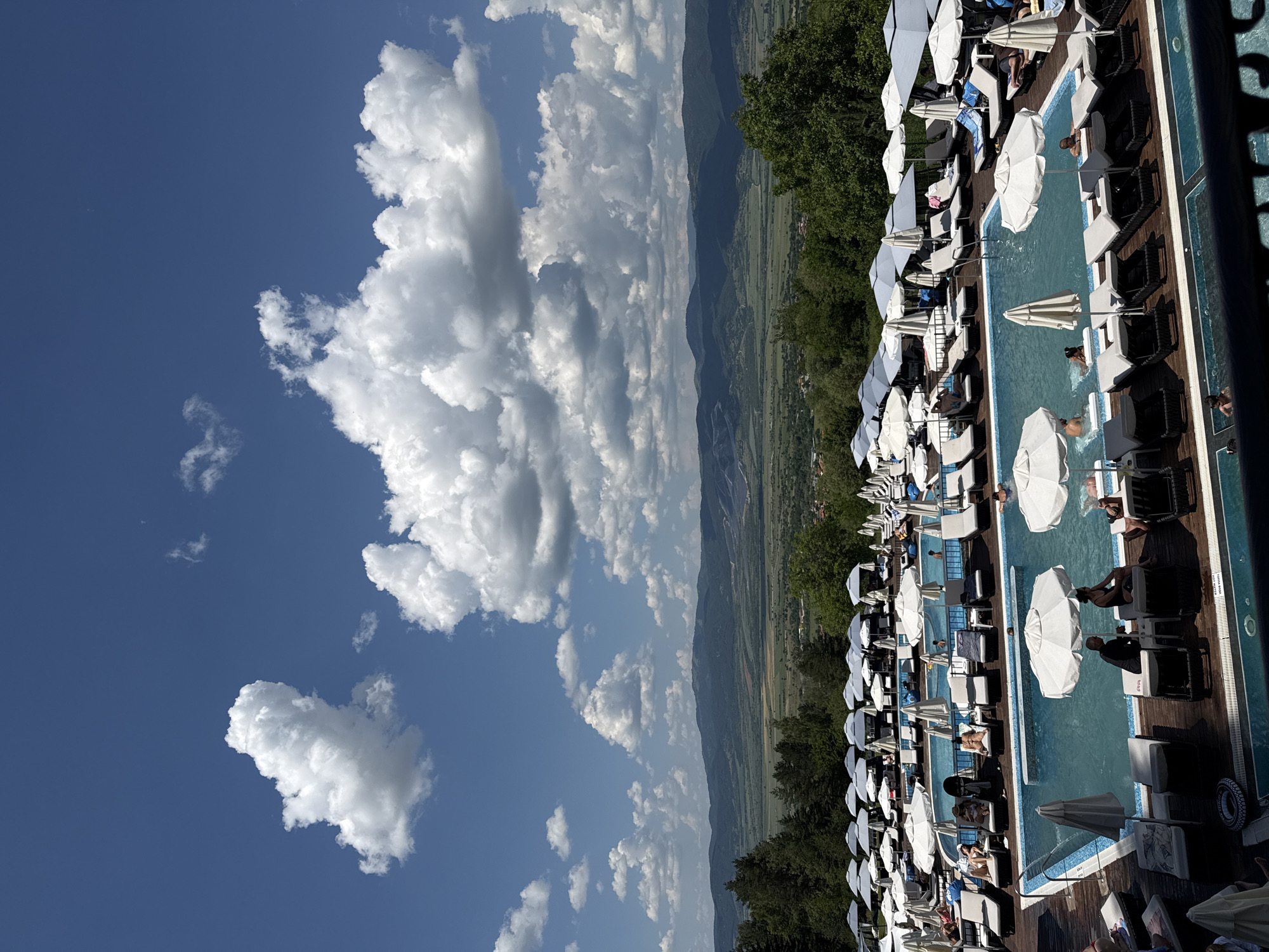 The image depicts a resort with multiple swimming pools and lounge chairs under white umbrellas, set against a backdrop of rolling green hills and a cloudy blue sky. In the foreground, people are seen relaxing on lounge chairs and swimming in the pools. The resort appears to be located in Sapareva Banya, Bulgaria, a town known for its hot springs and natural beauty. The scene conveys a sense of leisure and relaxation, with the clear blue water of the pools and the lush greenery of the surrounding landscape creating a tranquil atmosphere. The mountainous terrain in the distance adds to the picturesque scenery.
