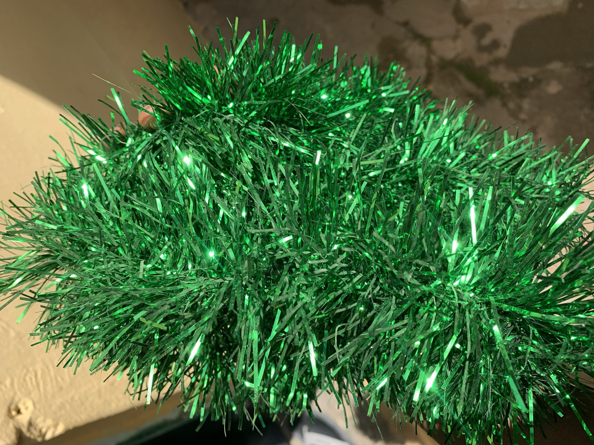 The image shows a close-up of green tinsel garland. The garland is composed of many thin, shiny strands of green material, which reflect light and give it a festive appearance. Some of the strands appear to have small, integrated lights that are emitting a green glow. The garland is bunched together, creating a dense and textured look. In the background, a blurred, light-colored wall or surface is visible, as well as a dark, indistinct area. A portion of a person's hand, with fingers, is visible through the tinsel in the upper left quadrant of the image. The lighting suggests that the photo was taken in artificial light, possibly indoors or at night. There is no visible text in the image. The context of Rafin Par, Nigeria does not provide specific visual cues in this image, as the subject is a decorative item.
