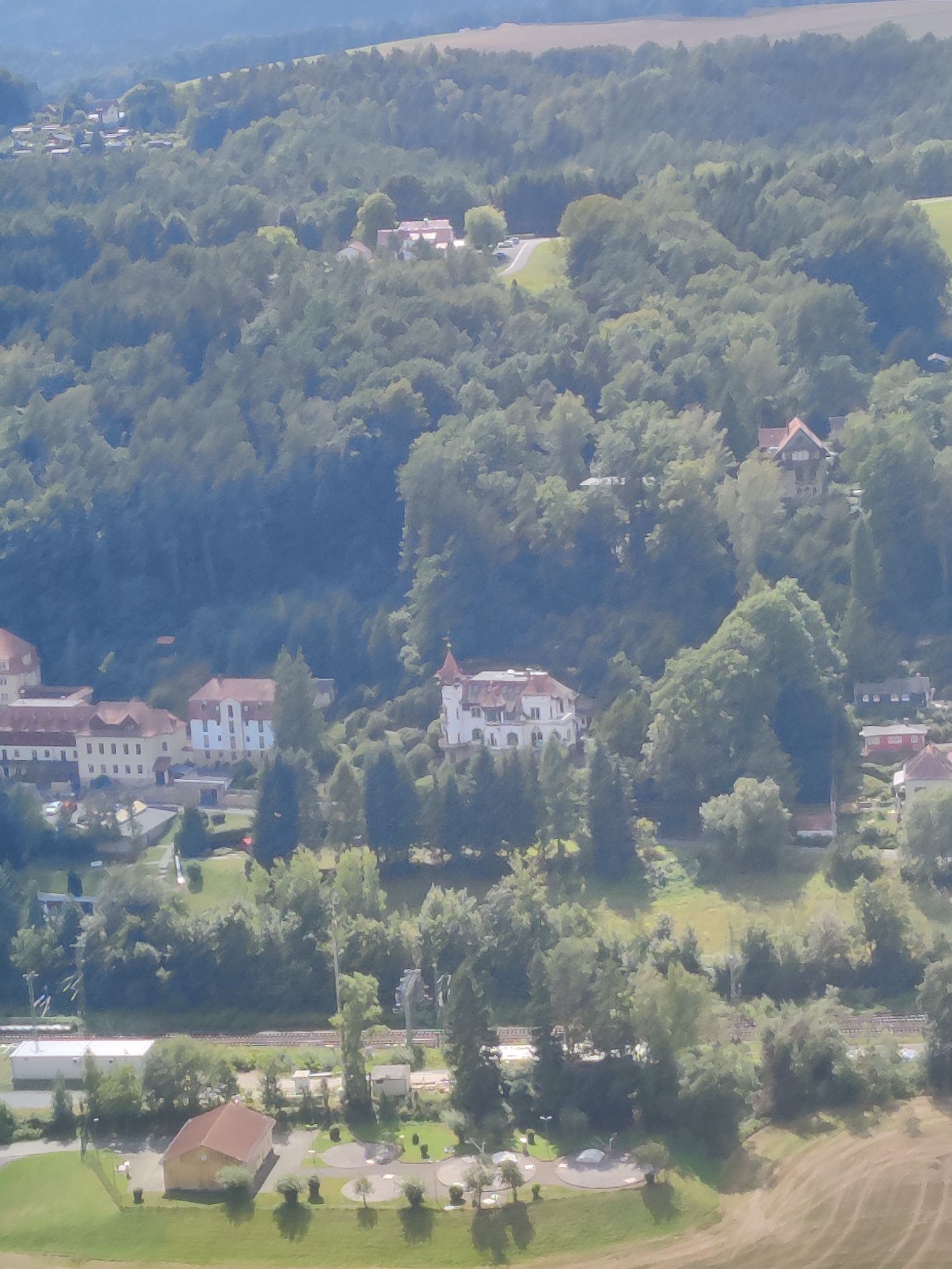 The image shows an aerial view of a town nestled among green hills and dense forests. In the foreground, a brown-roofed building, possibly a community center or a small school, is visible with several picnic tables and small trees surrounding it. A railway track runs horizontally across the middle ground, with a white building and some smaller structures adjacent to it.
Further into the distance, a cluster of buildings with red and white roofs, characteristic of German architecture, are scattered amidst the trees. One prominent building features a distinctive tower with a red roof. The overall impression is a serene, rural landscape with signs of human habitation integrated into the natural environment. The town of Lohmen, Germany, is known for its picturesque setting within the Saxon Switzerland National Park, which this image appears to depict.