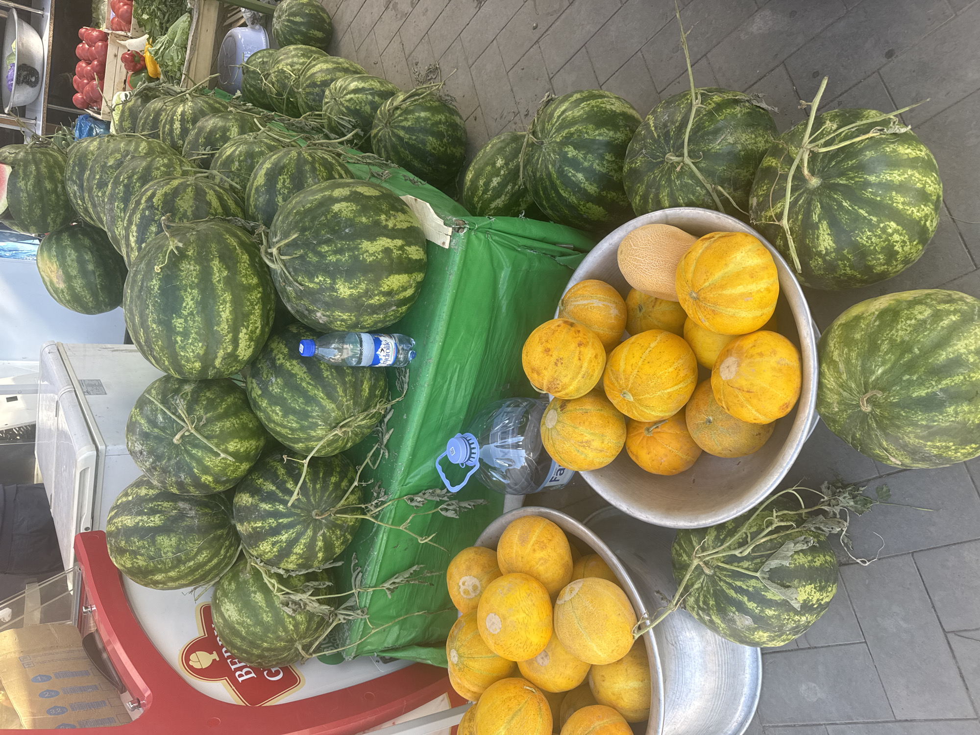 A market scene in Tashkent, Uzbekistan, displays a collection of watermelons and melons arranged for sale. Several green, striped watermelons are piled on a green tarp next to a white cart. Two silver-colored metal bowls are filled with small, yellow melons. Plastic bottles of water are also present. The setting appears to be outdoors, possibly a sidewalk, with gray paving stones visible. Other fruits and vegetables are barely seen in the background to the left.