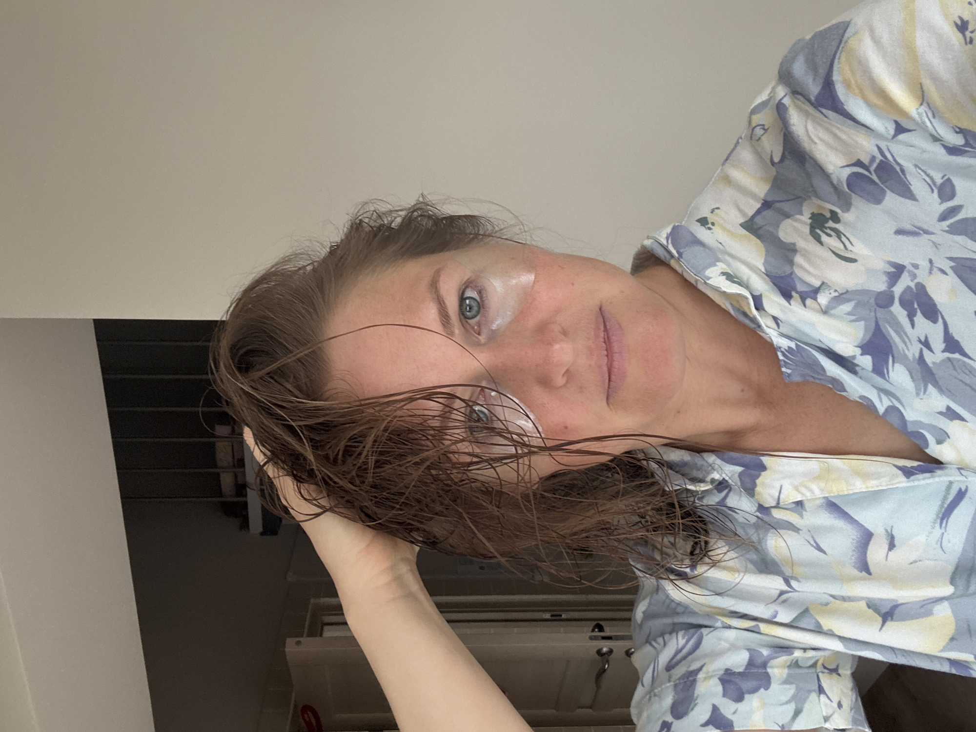 The image displays a woman in a close-up portrait, looking directly at the viewer. Her head is tilted, with her left hand lightly touching her wet, dark brown hair which falls across her forehead and face. She has light blue eyes and is wearing light pink lipstick. A small amount of white skincare product or cream is visible on her upper right cheek, near her temple. She is dressed in a light blue patterned shirt adorned with light yellow and purple-grey floral or abstract designs.
The background is simple and largely out of focus, featuring a light-colored wall or ceiling in the upper portion of the frame (when viewed upright). Below that, a darker, indistinct area suggests indoor furniture or cabinetry. The scene suggests a casual, personal moment, possibly post-shower or during a morning routine involving skincare. Her expression is calm and composed. There is no visible text. The indoor setting does not offer specific visual cues to confirm the location in Chaikovsky, Russia.