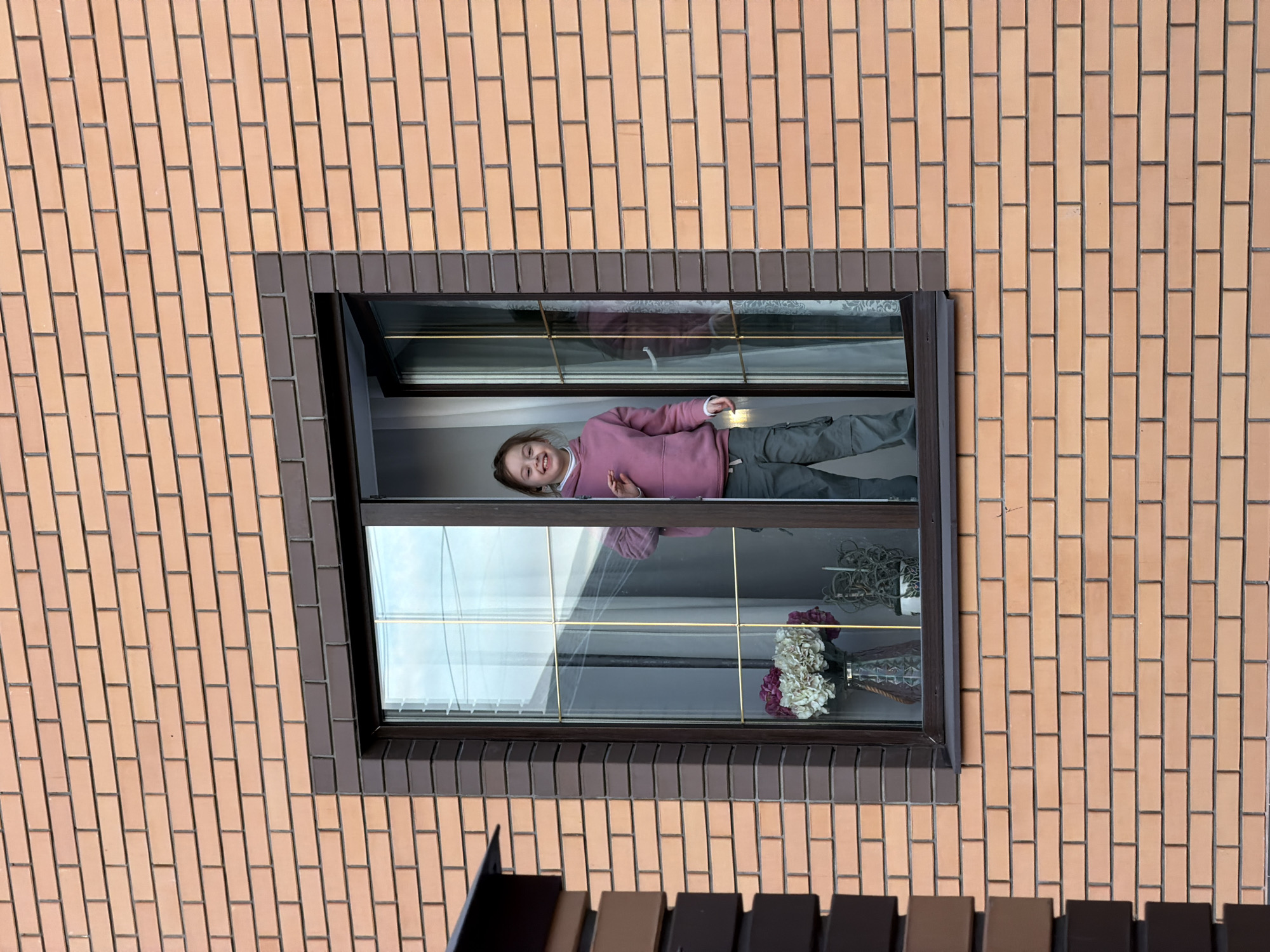 The image captures a section of a brick building exterior in Khimki, Russia, featuring a single window. The wall is constructed of light orange-brown bricks with dark mortar lines. The window has a dark brown frame; the left pane is fixed, while the right pane is a casement window partially open inwards. Through the left pane, interior details include a clear glass vase holding a bouquet of white and purple artificial hydrangeas, another potted plant, and white curtains. A young girl, appearing to be approximately 6-10 years old with brown hair, stands within the open right window section. She is smiling broadly, facing towards the viewer, and wears a pink hooded sweatshirt and olive-green cargo pants. Her right hand is slightly raised with the palm open, as if in a gesture. To the left of the window, a partial view of an architectural element, composed of dark and light brown brick-like blocks, is visible.
