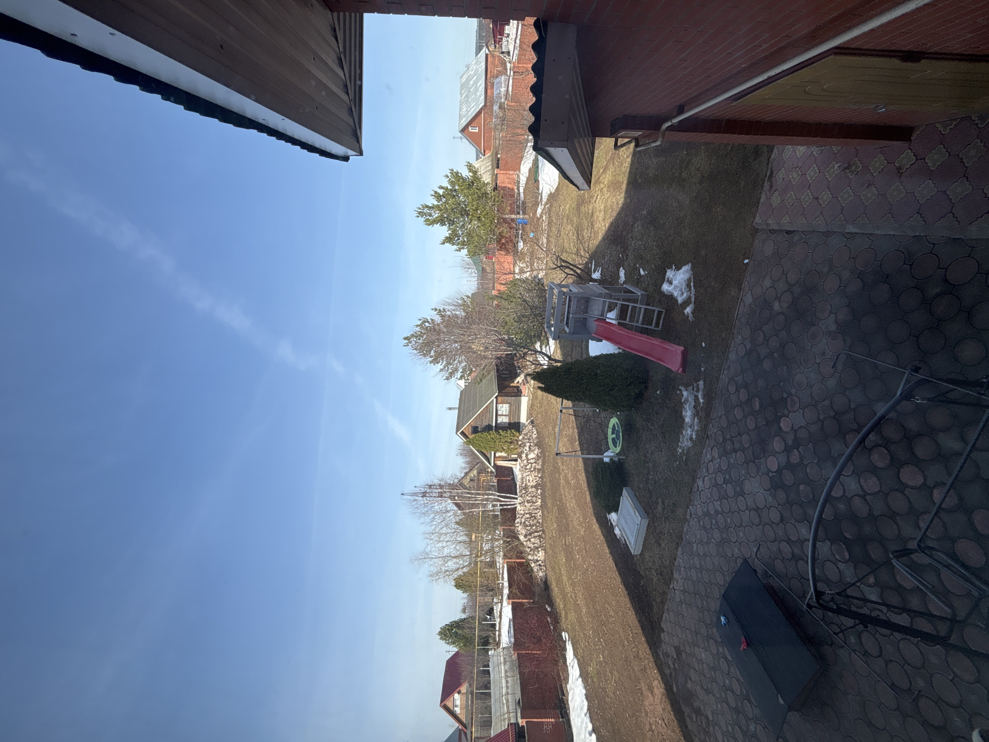 The image depicts a view from a building, likely a house, in Ферма, Russia, looking out over a residential area. The sky is clear blue with a few wispy clouds. The ground is covered in grass, with patches of melting snow in some areas. There are several houses visible in the distance. In the immediate foreground, there is a children's playground set, a slide, and a couple of trees. The camera is angled upward towards the sky. A fragment of the building from which the picture is taken is visible at the top and sides. The foreground floor surface is paved.