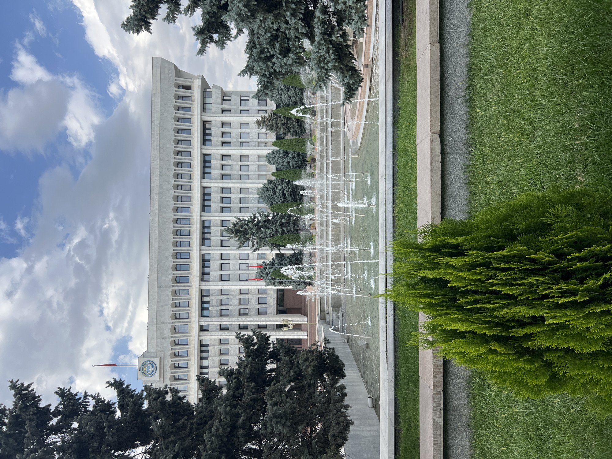 The image captures a frontal view of the White House, a government building in Bishkek, Kyrgyzstan, characterized by its light-colored stone facade and a distinctive cantilevered upper story. In the foreground, a tiered fountain with multiple water jets is active, with water spraying upwards. Lush green grass and a neatly trimmed evergreen bush are visible in the immediate foreground, separated from the fountain area by a concrete planter. Large evergreen trees frame the sides of the building and the fountain. Two red flags are prominently displayed on poles in front of the building's entrance, and a national emblem with a flag is visible on the building's rooftop. The sky above is partly cloudy.