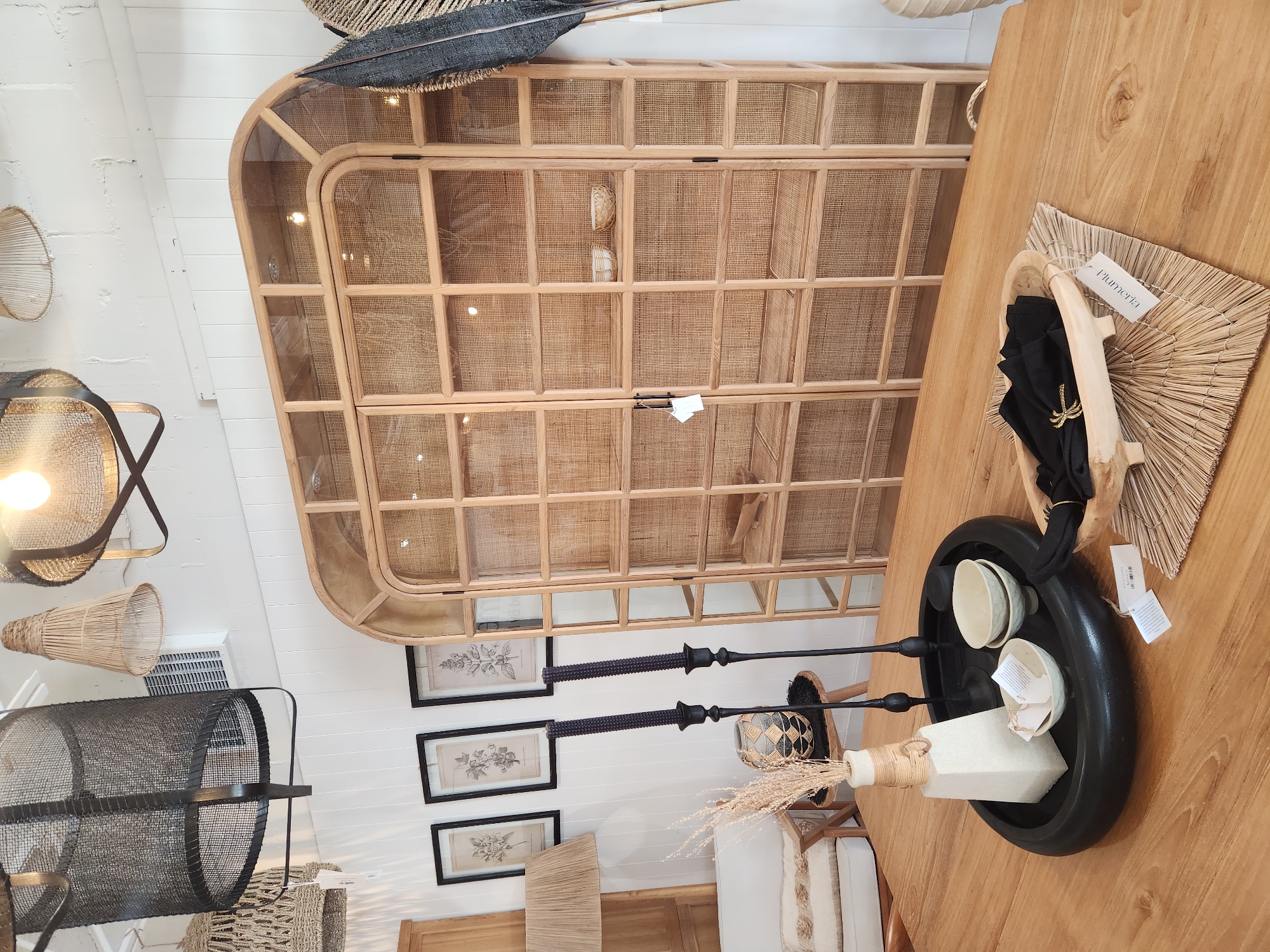 The image displays a retail store interior, likely a home decor shop, set in Maitland, Florida. The focal point is a large, rounded, glass-fronted cabinet with a cane-style insert, crafted from light-colored wood. Inside the cabinet are several shelves displaying small decorative bowls. In the foreground, a wooden table features a black oval tray containing a rectangular off-white vase with dried reeds, two black candlesticks, and three small ceramic bowls. To the right of the tray, a wooden bowl filled with black napkins rests on a woven placemat. In the background, a white wall is adorned with three framed botanical prints. Several pendant light fixtures, with woven and metal shades, hang from the ceiling. A portion of a white sofa is visible to the left. The overall scene suggests a curated display of natural and handcrafted home furnishings.