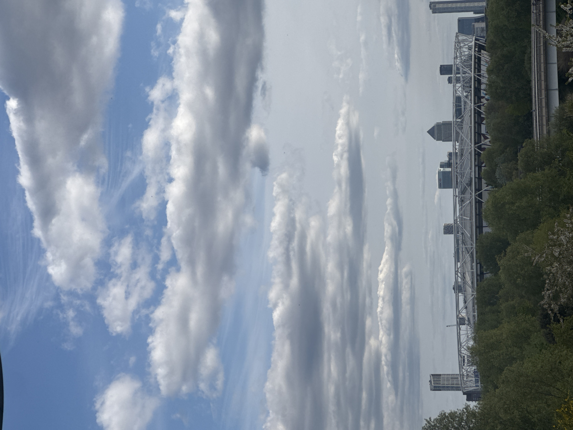 The image is a vertical shot of the London cityscape under a partly cloudy sky. The clouds are predominantly white and gray, with varying densities and formations. A portion of the city skyline is visible, including the Canary Wharf financial district. The infrastructure of the area features a white pedestrian bridge spanning a body of water. The landscape includes trees along the bridge and waterfront. The scene is taken during daylight hours.