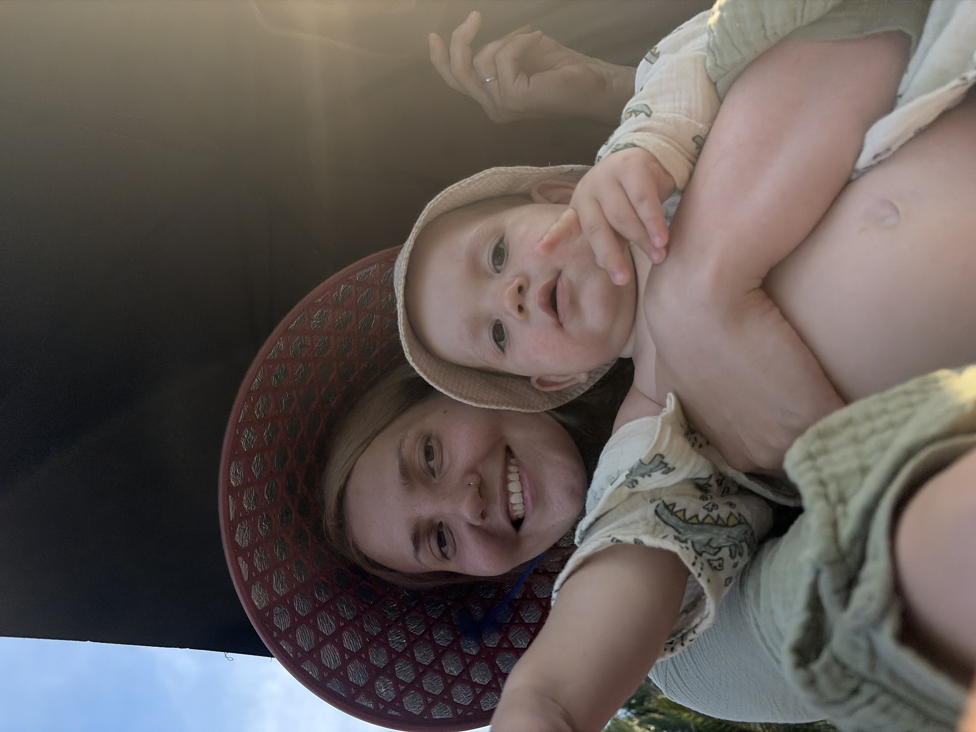 This low-angle portrait captures an adult female holding a baby or toddler in what appears to be a sunny outdoor setting, likely in Đà Nẵng, Vietnam. The adult, positioned in the lower part of the frame, has light hair and a nose piercing, and wears a wide-brimmed hat with a distinctive red woven pattern. She smiles warmly and broadly at the camera. Her left hand, wearing a ring, is visible near the baby's head.
The baby, held securely by the adult, wears a light-colored bucket hat and a pale garment adorned with a charming crocodile or alligator pattern. The baby gazes towards the camera with wide, curious eyes and a slightly open mouth, displaying a neutral yet engaged expression. Bare legs and a visible belly button on the baby suggest light attire suitable for warm weather.
The setting includes a dark, flat overhead surface that covers the majority of the upper frame, possibly an awning, umbrella, or boat canopy, providing shade. A strip of clear blue sky is visible at the very bottom of the image, confirming the outdoor location. Bright sunlight or lens flare streaks across the upper-middle section, indicating a sunny day, perhaps during the morning or late afternoon. The subjects appear to be posing for a casual photo, with a joyful and affectionate mood conveyed by the adult's smile. There is no visible text in the image.