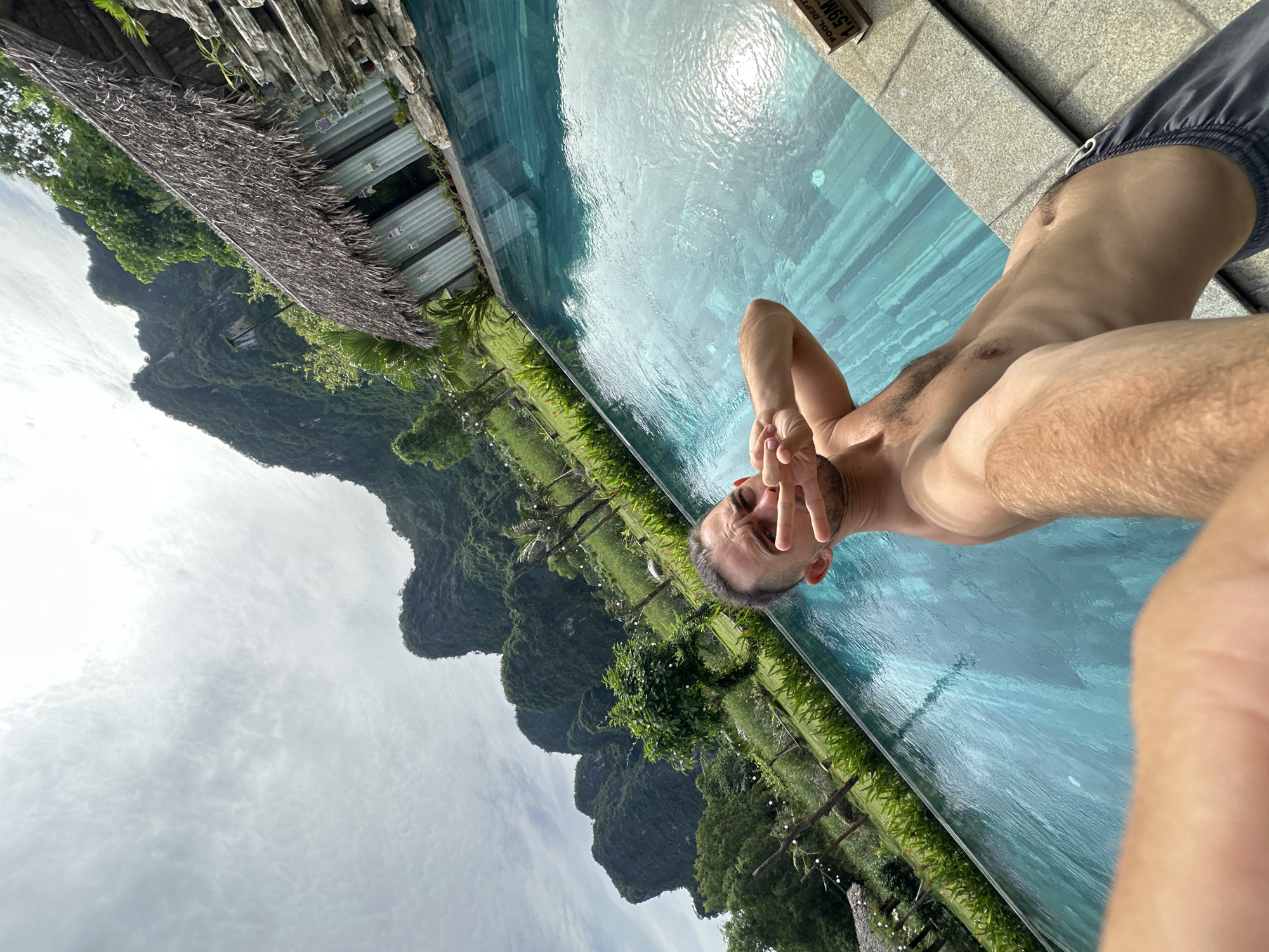 A shirtless man with dark hair and a goatee poses for a selfie by an outdoor swimming pool. He is wearing dark swimming shorts and holds up two fingers in a peace sign towards the camera. The pool is filled with turquoise water and has a tiled edge. In the background, lush green mountains rise dramatically against a cloudy sky. A thatched-roof structure with metal siding is visible next to the pool, and beyond that, a landscape of trees and vegetation. A sign indicating "POOL DEPTH 1.59M" is visible on the pool edge. The scene appears to be in Nam Hoa Lư Ward, Vietnam, known for its karst topography and natural beauty.