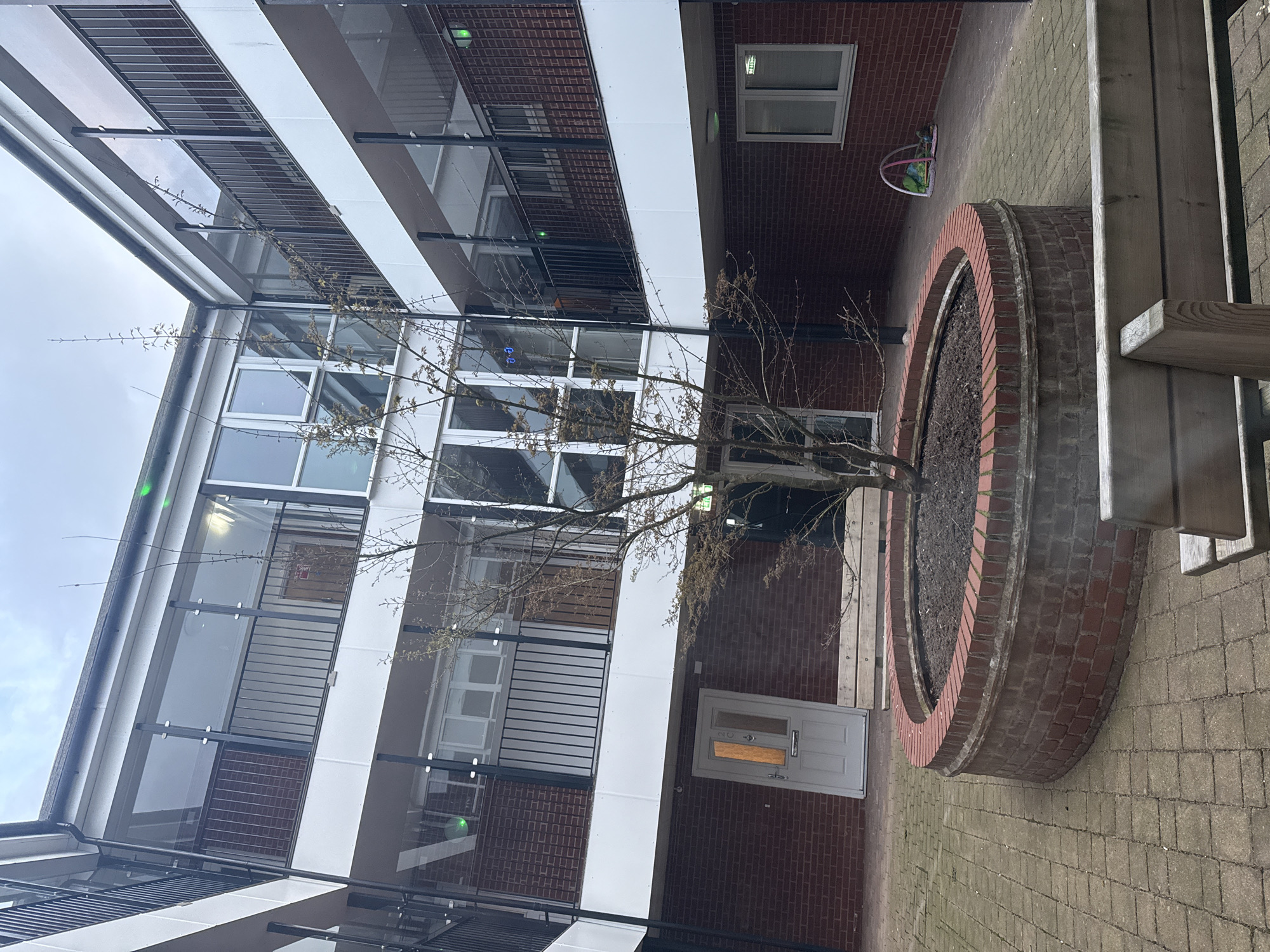 The image depicts an exterior courtyard or enclosed space within a building complex in Gorleston-on-Sea, United Kingdom. The scene features a central circular brick planter containing a bare tree. The surrounding architecture consists of a multi-story building with a mix of brick and white paneling, interspersed with windows and metal balconies. A ground-level doorway is visible in the brick facade. A section of a wooden bench sits on the right edge of the frame, bordering a paved area. The sky overhead appears overcast, with glimpses of it visible at the top of the frame. No people are visible in the scene.
