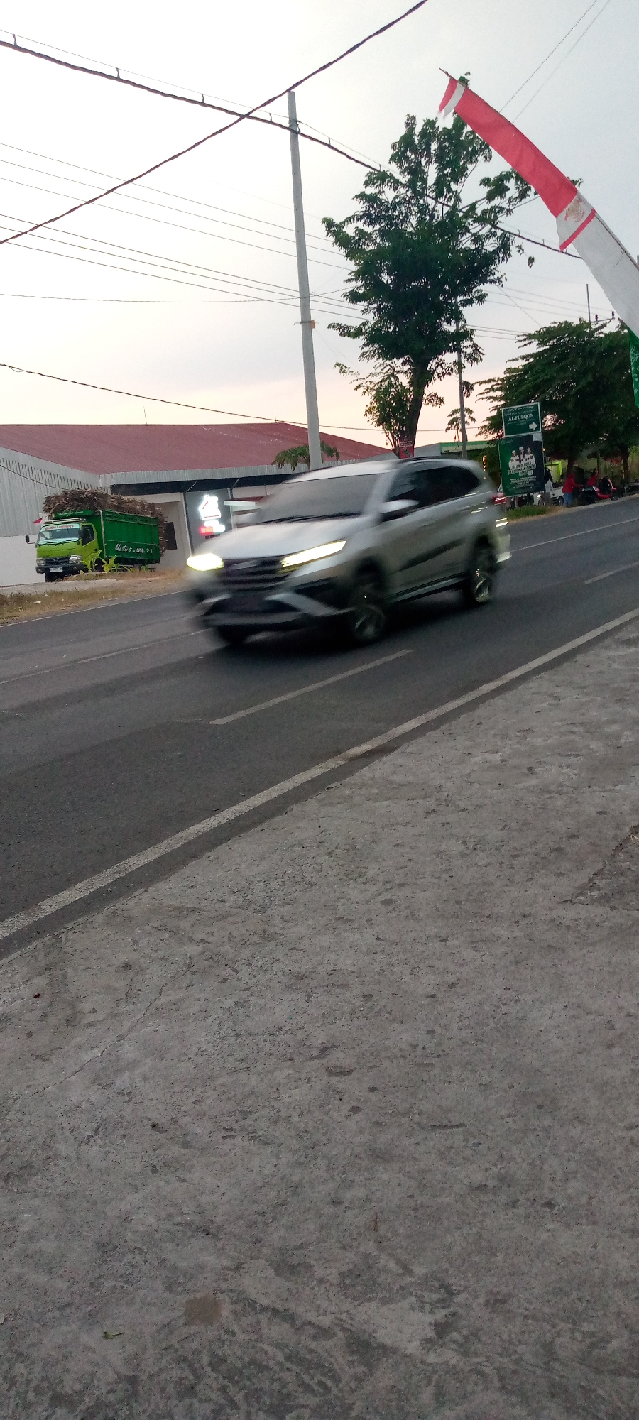 In the city of Sumberwono, Indonesia, a silver car is captured in motion, its headlights illuminated, on a paved road. The car is the primary subject, its form blurred due to its speed. To the left of the road, a green truck is parked, laden with what appears to be logs or branches. Behind the truck, a building with a red roof and white facade is visible. Further in the background, trees line the side of the road. An Indonesian flag, red and white, is seen fluttering in the upper right portion of the image. The sky is overcast, suggesting either early morning or late evening light. The road itself is marked with white lines and transitions to a concrete surface on the right side of the frame. Power lines are strung across the sky, supported by utility poles.