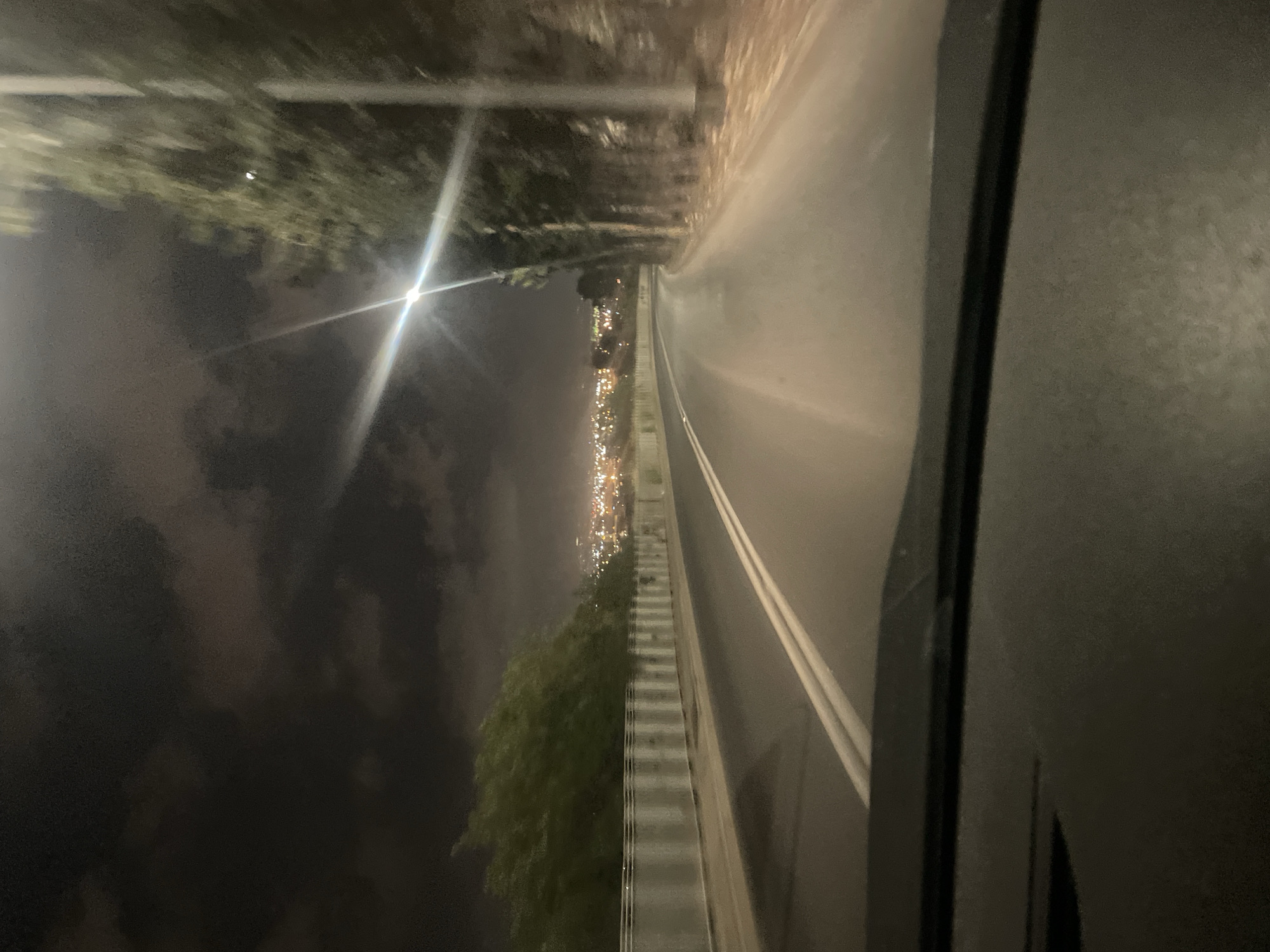 The image captures a nighttime view from inside a vehicle driving along a road that curves to the right. The road has a solid white barrier on the left, topped with vertical posts and a metal railing. Beyond the barrier, lush green trees line the side of the road. On the right side, a row of tall, slender trees creates a dark silhouette against the night sky. In the distance, a cityscape with numerous lights illuminates the horizon, suggesting the presence of a populated area. The sky is cloudy and dark, with a bright streetlight casting a diagonal beam of light across the scene. The foreground shows the dashboard of the vehicle, a dark and reflective surface. The overall scene depicts a drive through a semi-urban or suburban area at night, with the distant city lights providing a sense of scale and destination. The location is specified as a road in Haifa, Israel.