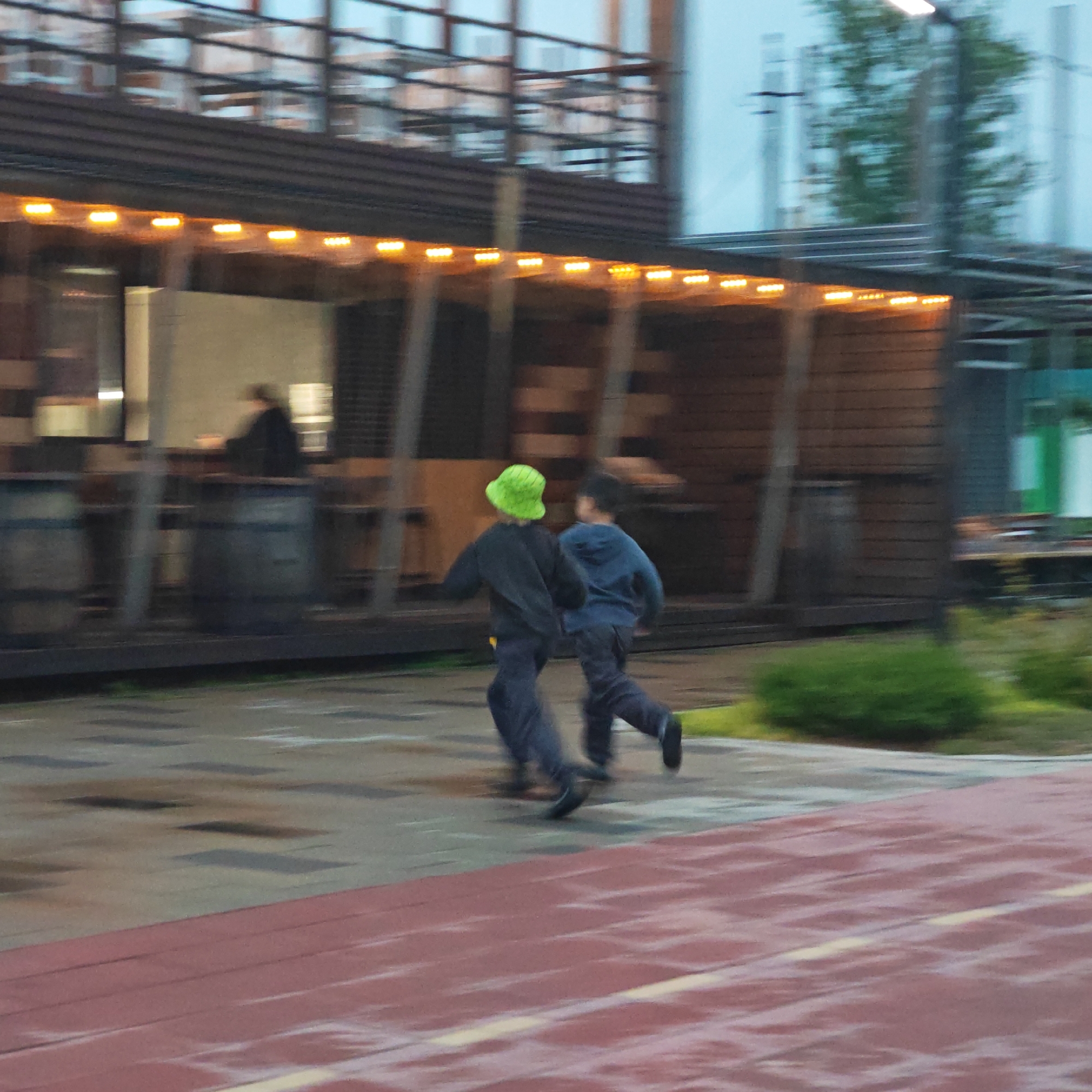 Two children are running on a red track in the городское поселение Щелкино, Ukraine. One child is wearing a green hat and a dark jacket, while the other is wearing a blue jacket. They are running past a building with lights and barrels. The ground appears wet.