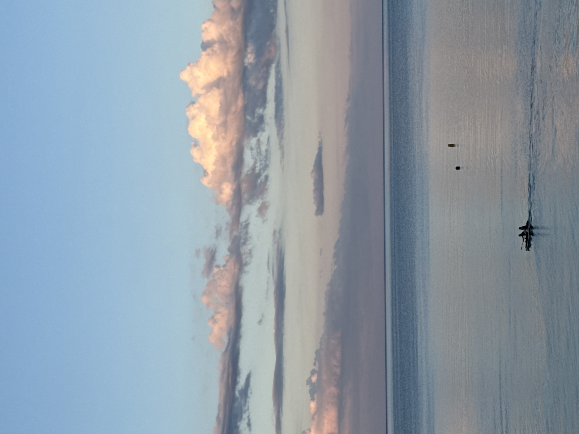 The image shows a serene seascape with a pale blue sky and soft pink clouds at sunset. The calm, reflective water stretches to the horizon, meeting the sky in a seamless blend of blues and purples. In the mid-ground, a small, dark-colored raft with two silhouetted figures paddling is visible. Further out in the water, there are two buoys, one dark and spherical, the other a cylindrical, light-colored object. The overall impression is one of tranquility and the peaceful end of a day.