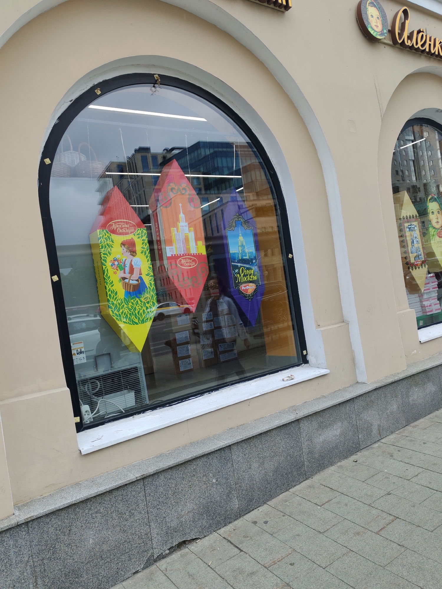This image captures a storefront in Moscow, Russia, featuring two large arched display windows set into a light beige building facade with white architectural trim. The foreground shows a sidewalk paved with grey concrete pavers and a low wall clad in grey granite slabs.
Inside the left display window, three large, colorful packages, shaped like elongated pyramids or cones, are prominently displayed. The left-most package is yellow and green, illustrated with a girl holding flowers and a basket, bearing the text "Красный Октябрь" (Krasny Oktyabr) and "конфеты" (sweets). The middle package is red and orange, depicting a tall Moscow skyscraper and also displaying "Красный Октябрь." The right-most package is purple, featuring a cityscape with a prominent building (likely Moscow State University) and the text "Огни Москвы" (Lights of Moscow) and "РотФронт" (RotFront). Reflections in the window show modern buildings, a yellow taxi, and an air conditioning unit within the store.
Above the right window, a round sign with the iconic face of the "Алёнка" (Alyonka) girl, a famous Russian chocolate brand, is visible next to the Cyrillic lettering for "Алёнка." The second display window to the right is partially visible, also containing reflections and possibly more product displays.
The scene depicts a commercial establishment, likely a confectionery or chocolate shop given the prominent branding of well-known Russian sweet manufacturers "Krasny Oktyabr," "RotFront," and "Alyonka." It appears to be daytime, possibly overcast, with no direct sunlight. No people are directly visible, and no specific activity other than product display is taking place.