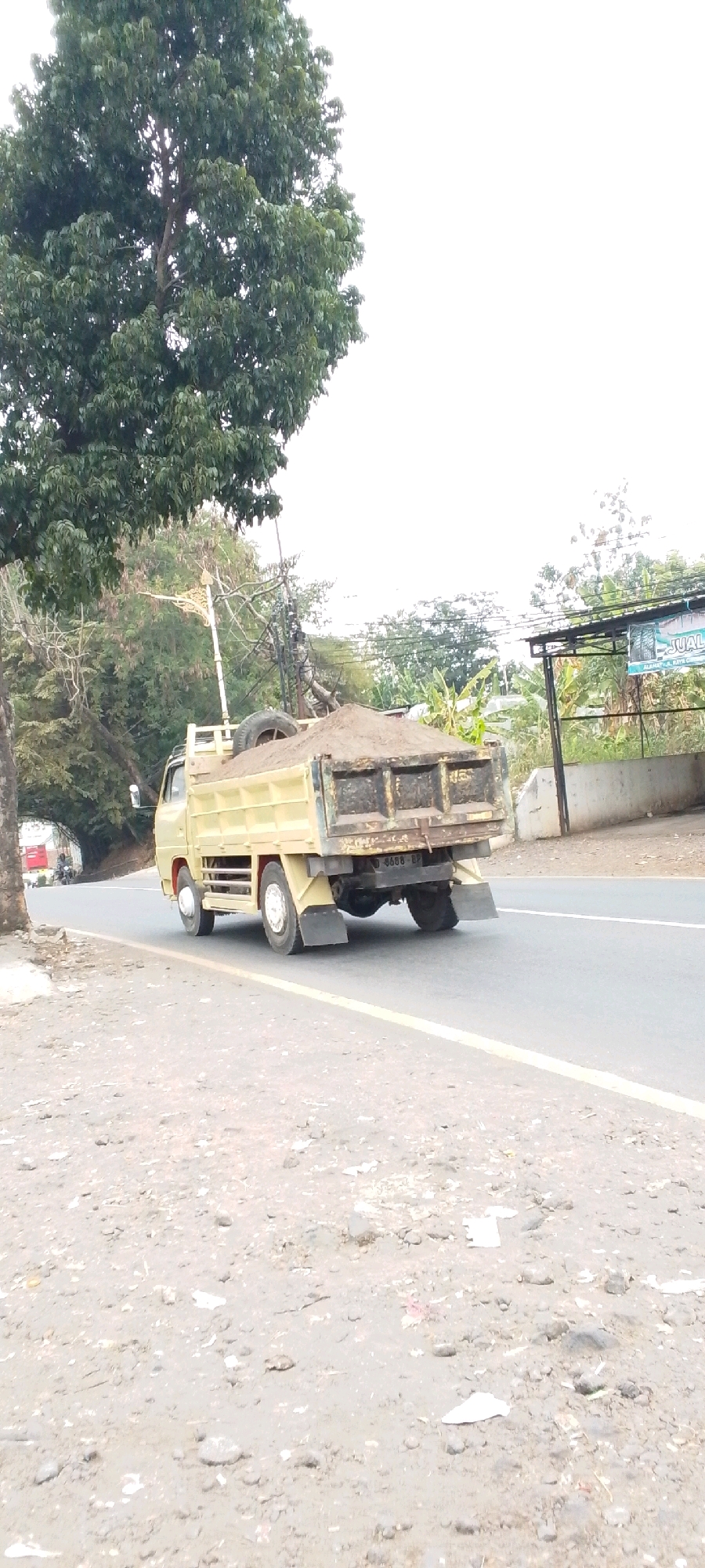 A yellow dump truck is traveling on a paved road in Cilaku, Indonesia. The truck is loaded with sand. The truck has a license plate that reads "D 6686 BP". A large tree with green leaves is on the left side of the image, and power lines are visible in the background. The sky is overcast. The road is lined with trees and vegetation. There are some signs in the background. The foreground is a dirt and gravel shoulder of the road.