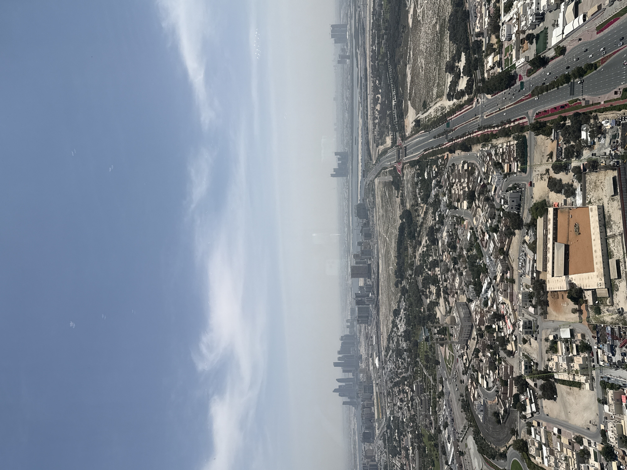 This image provides an expansive aerial view of a modern urban landscape, likely Dubai, under a daytime sky. The perspective is from a significant height, looking down upon a vast city stretching towards a hazy horizon.
The ground portion of the scene features a complex network of multi-lane highways and roads, with numerous vehicles indicating active traffic. These arteries cut through a mix of developed and undeveloped areas. Residential and commercial buildings, ranging from low-rise structures with light-colored facades to clusters of modern high-rises in the middle distance, populate the urban fabric. Interspersed among the buildings and roads are patches of green vegetation, alongside large sections of sandy, undeveloped terrain, characteristic of a desert environment. A linear body of water, possibly a canal or creek, is visible in the middle ground, snaking through the urban sprawl.
The sky, occupying a significant portion of the image, is predominantly a light, clear blue with scattered, wispy white clouds visible on the right. Several small, translucent, bubble-like specks are noticeable in the upper left section of the sky, likely reflections or debris on the camera lens.
The time of day appears to be mid-morning or early afternoon, with bright, even illumination across the landscape. Atmospheric haze is present in the distance, obscuring the clarity of the furthest buildings and the horizon, contributing to a muted appearance of the distant urban areas. The image captures the scale and ongoing development of a bustling metropolis in an arid region. No discernible text is visible within the frame.
