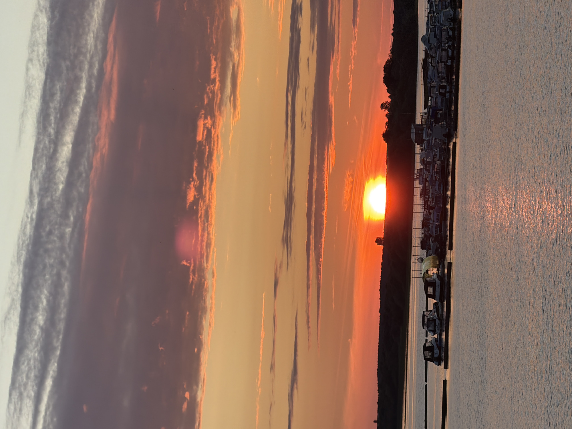 The image shows a serene waterscape at sunset. The sun, a brilliant orb of light, is setting over a body of water, casting a warm, golden glow across the scene. The sky is a canvas of vibrant colors, with streaks of orange, pink, and purple clouds painting a dramatic backdrop. The sun's reflection shimmers on the rippling surface of the water, creating a mesmerizing effect.
Along the shoreline, a marina is visible, with numerous boats docked and awaiting their next journey. The boats, mostly small to medium-sized, are neatly arranged, their silhouettes adding a sense of order to the tranquil environment. A thin line of trees forms a dark silhouette against the fiery sky, marking the edge of the land.
The time of day is clearly sunset, indicated by the low angle of the sun and the rich, warm hues in the sky and water. The weather appears to be clear and calm, with no signs of wind or precipitation. The overall mood is one of peace and tranquility, inviting viewers to soak in the beauty of nature's spectacle. While no people are explicitly visible, the presence of the marina suggests human activity and leisure associated with boating. The location context of Solikamsk, Russia, places this scene in a specific geographical setting, hinting at the natural beauty of the region. There is no visible text in the image.