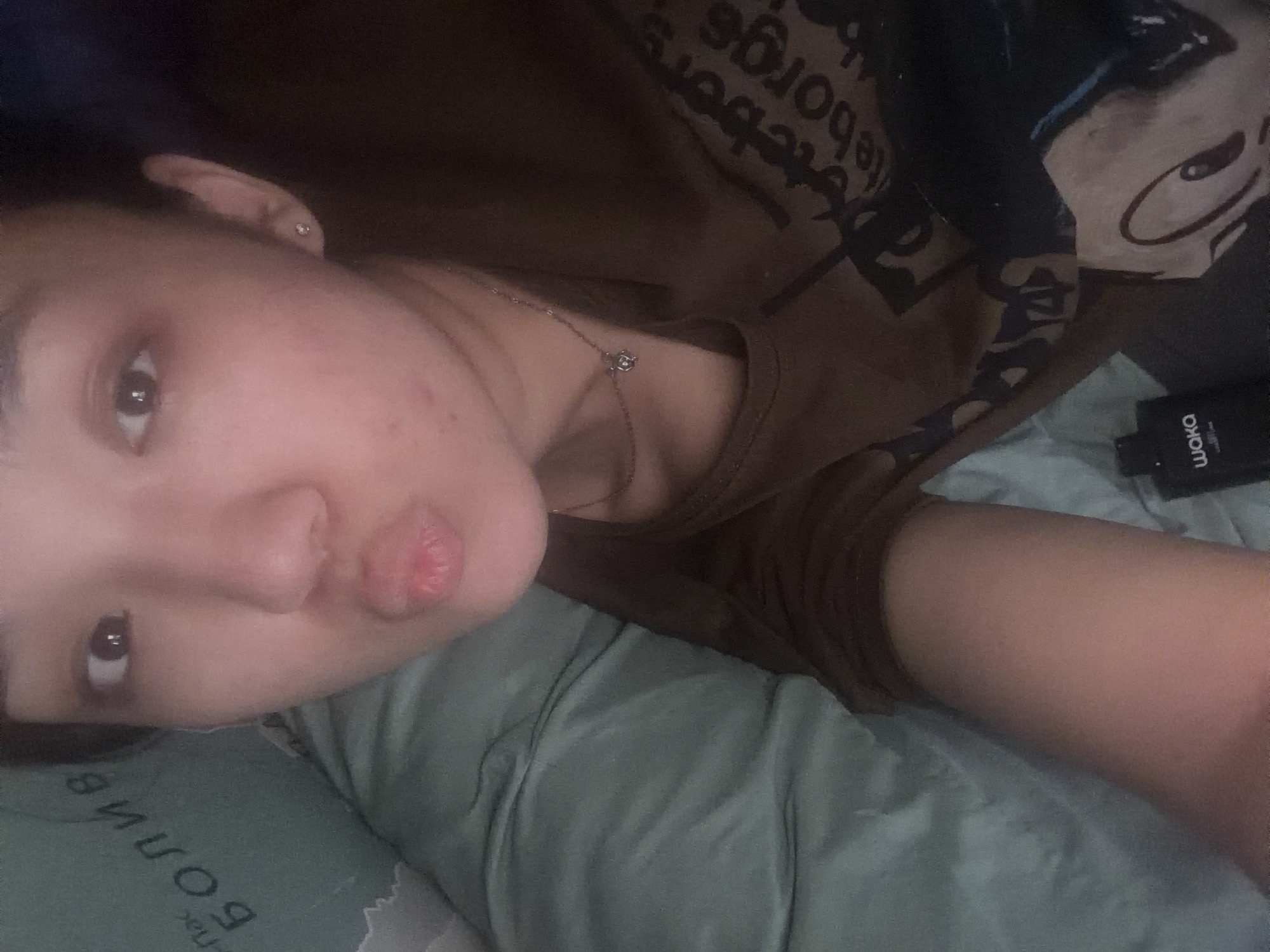 A young East Asian female is lying down and looking at the camera, puckering her lips. She is wearing a brown t-shirt with a graphic print and a delicate silver necklace. Her earlobes are pierced with small stud earrings. To her left, a teal-colored pillow with Russian text is visible. A black bottle with the word "waka" on it is placed near her chest. The background is a soft, rumpled fabric, suggesting a bed or sofa.