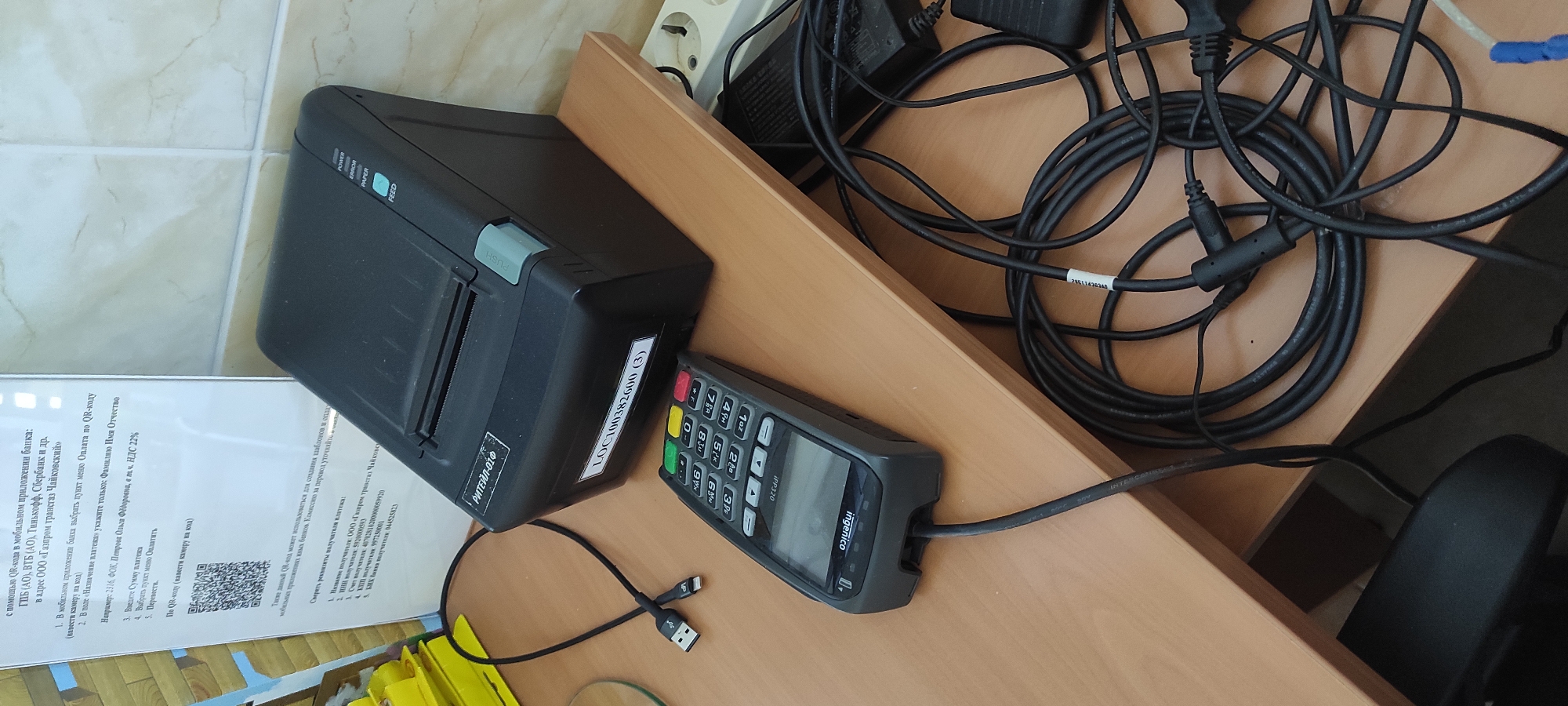 The image shows a desktop scene that appears to be in an office or a similar commercial setting. On the right side, a black thermal printer with a label "LOC100382600 (3)" is positioned on a wooden desk. To the left of the printer, a gray Ingenico IPP320 point-of-sale (POS) terminal is also on the desk, connected by a black cable. In the background, there's a white tiled wall and a beige wall. A power strip with multiple outlets is visible on the right side.
The foreground is dominated by a tangle of black cables, including USB cables and power cords, connected to the POS terminal and possibly other unseen devices. A USB-to-Lightning cable is also present.
On the left side of the image, a white piece of paper with text and a QR code is affixed to the wall or a bulletin board. The text appears to be instructions or information related to mobile payments and bank transactions, mentioning "QR-code," "mobile application," "bank," and specific financial institutions. The presence of a QR code suggests a digital payment system is in use.
The overall environment appears functional and somewhat cluttered due to the network of cables. There are no people visible in the image. The scene is likely an indoor workspace equipped for financial transactions. The mention of "Noviy, Russia" is not directly supported by visual elements in the image itself, but if this context is provided, it indicates the geographical location of this scene.