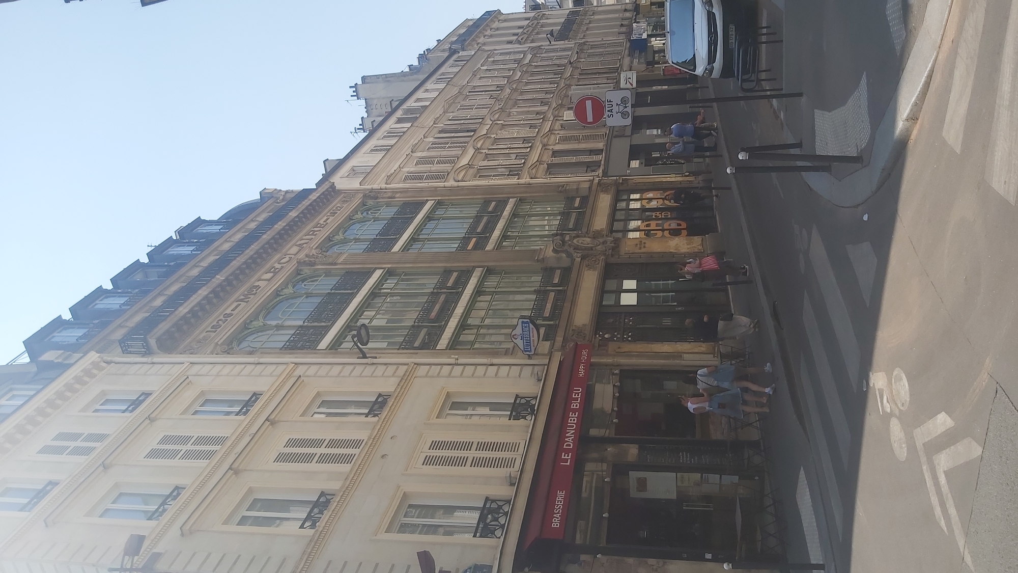The image captures a street scene in Paris, France, on a clear day. The perspective is from a low angle, looking up at the architecture and down at the street.
On the left side of the image, a tall, cream-colored building with multiple stories dominates the frame. This building features numerous windows with white shutters, organized in a grid pattern. Below these windows, a red awning with "BRASSERIE LE DANUBE BLEU HAPPY HOURS" is prominently displayed, indicating a restaurant or cafe. A few people are visible on the sidewalk in front of this establishment, some appearing to be customers.
Adjacent to the cream-colored building is a taller, darker building with more elaborate architectural details. This building exhibits a series of large, arched windows on its upper floors, framed by decorative stonework. The inscription "1826-Mme LE CLAIRE-1912" is visible across the facade, suggesting historical significance. A blue sign with the word "LUTTERBACH" is also present.
Further down the street, to the right, a series of narrower, multi-story buildings with shutters line the road. A white van is parked on the right side, partially visible. A pedestrian crossing is marked on the asphalt, and a bicycle symbol is painted on the road surface. Traffic signs, including a circular red sign with a horizontal bar and a sign with a bicycle symbol, are also visible, indicating traffic regulations.
The environment appears to be an urban setting with clear skies. Shadows are cast on the street, suggesting the sun is positioned to the right and slightly behind the viewer. The overall impression is a bustling street with a mix of historical and modern elements.