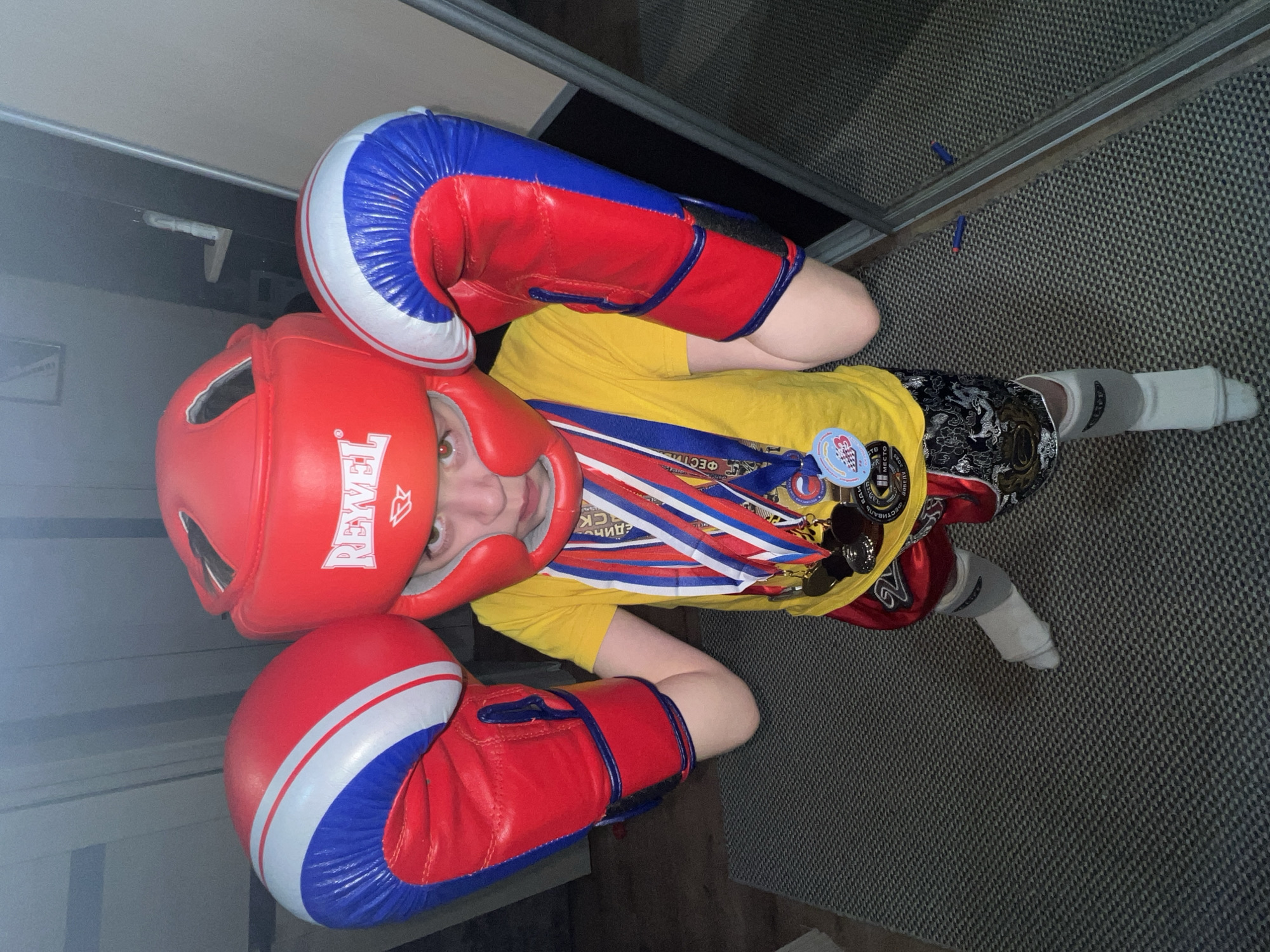 A child is lying on the floor, wearing a red boxing head guard with the word "REVEL" on the side and red and blue boxing gloves. They are also wearing a yellow t-shirt with multiple medals and pins attached. Their legs are partially visible, with one foot covered in a white sock or wrap. The floor appears to be carpeted in a neutral tone. A sliding closet door is visible in the background. This is unlikely to be located in the city of Noviy, Russia.