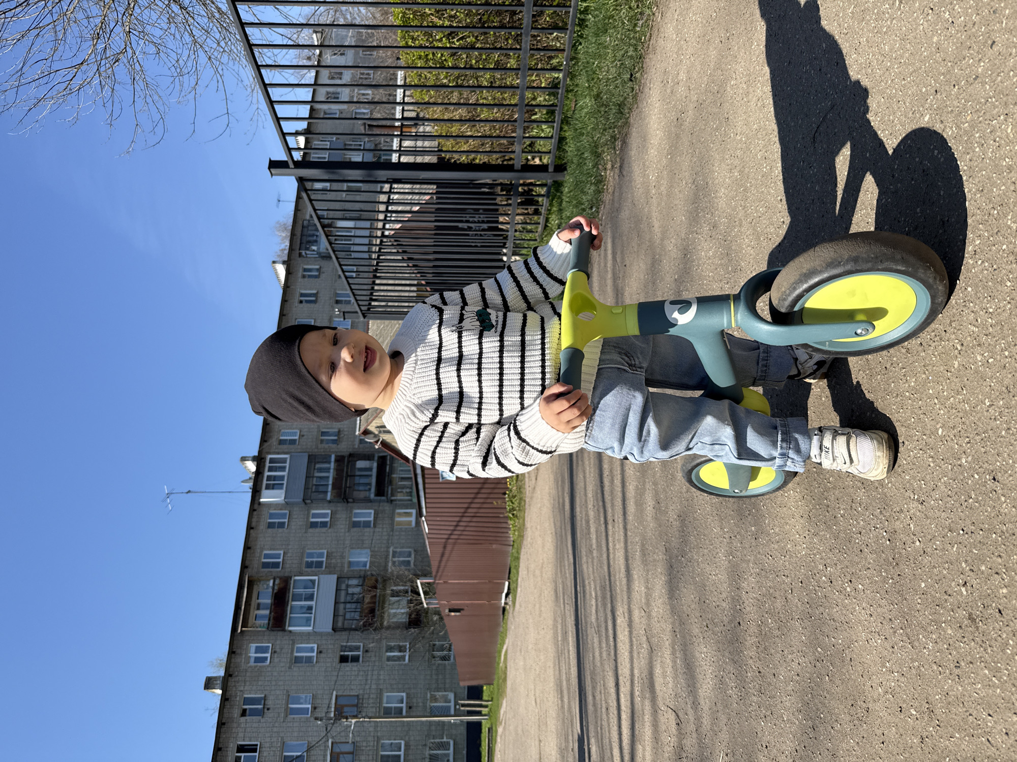 A young child on a balance bike is the central figure of the image, positioned on a concrete surface. The child is wearing a striped sweater, jeans, white sneakers, and a dark beanie. The balance bike is green and yellow.
The background includes a multi-story residential building with numerous windows. A fence runs along the middle ground, with some greenery visible behind it. The sky is blue. The environment appears to be urban. The city is not specified.