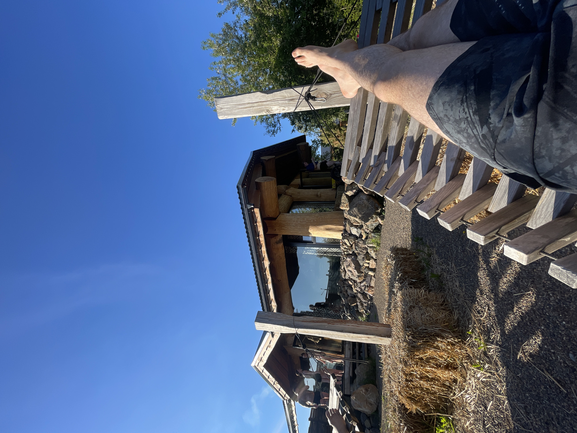 The image, taken in Дрокино, Russia, captures a bright, sunny outdoor scene with a focus on leisure. In the foreground, the bare feet and lower legs of a person are propped up on a slatted wooden bench or lounger. The person is wearing dark, patterned shorts.
In the midground, a rustic wooden cabin or sauna stands under a clear, vibrant blue sky. The building is constructed with large, rough-hewn logs and features a dark roof. A large window on the facade reflects the sky and parts of the surroundings. Vague figures of at least two other people are visible inside or near the building, one appearing to be seated and another partially visible, possibly in dark swimwear. A sturdy wooden post, secured with ropes, is present in the immediate foreground, adjacent to the building.
To the right of the wooden structure, a bale of hay or straw rests on gravelly ground, suggesting a rural or natural setting. Green foliage from a tree is visible in the upper left portion of the image. Strong sunlight casts clear shadows, indicating it is daytime, likely late morning or afternoon. The overall impression is one of relaxation and enjoying the natural environment. No visible text is present in the image.