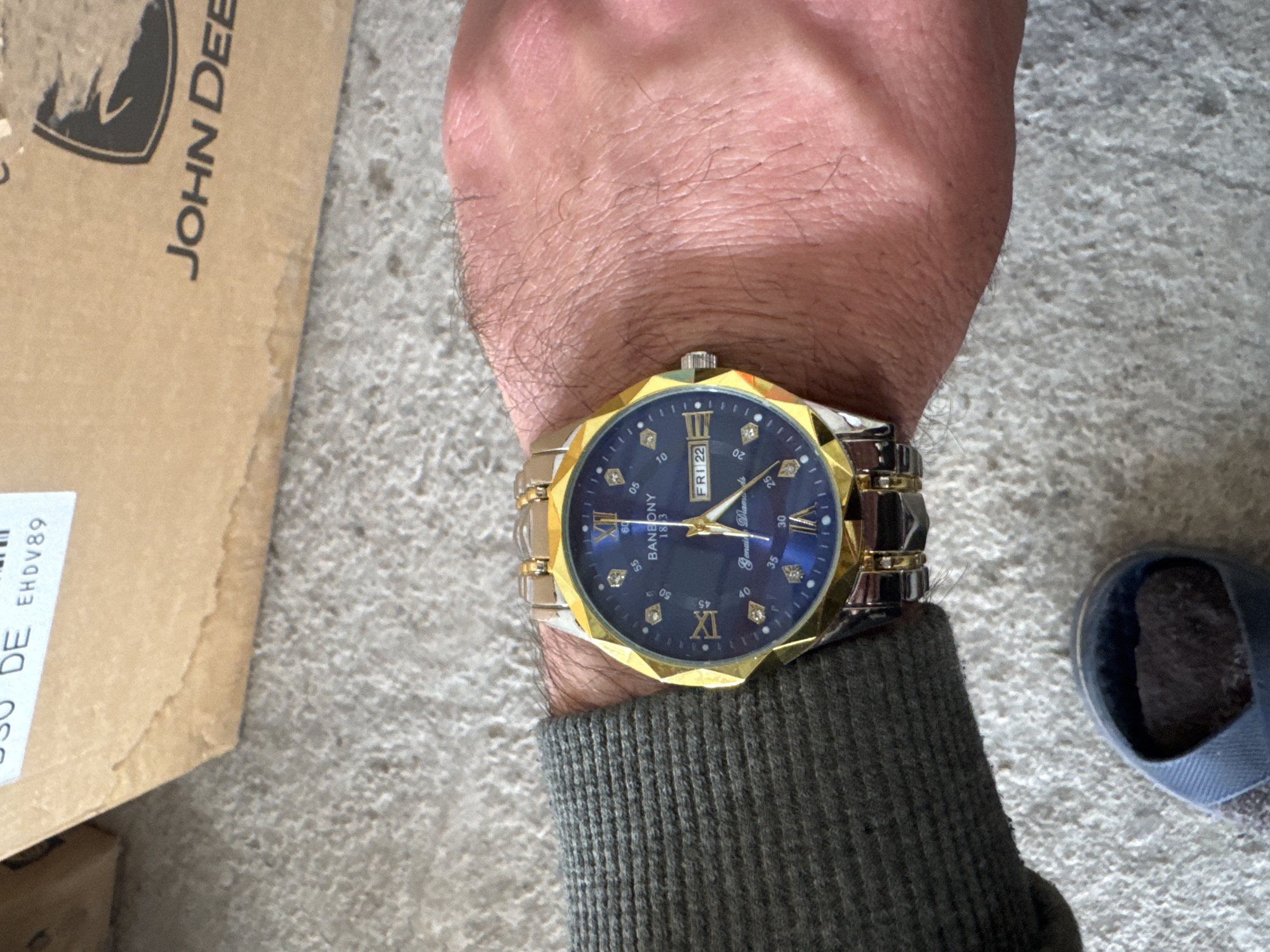 The image shows a person's wrist wearing a Baneony brand watch with a blue face, gold bezel, and silver and gold band. The watch displays "FRI 22" for the day and date. The wrist is surrounded by a dark gray sleeve. In the background, there is a portion of a cardboard box with "JOHN DEE" printed on it and a partial view of a blue slipper. The location of Sarnevo, Bulgaria, is not discernible from the image.