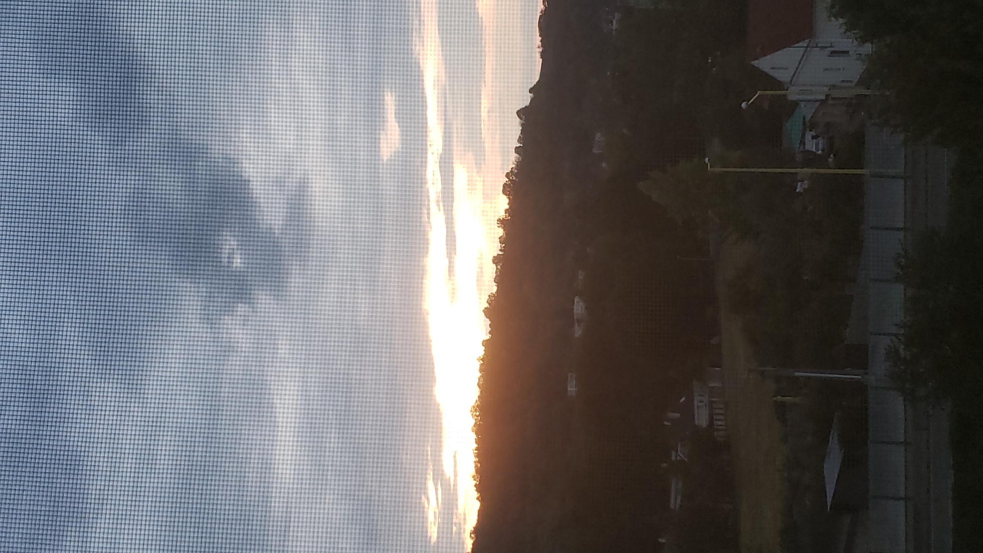 The image shows a sunset over a hilly landscape with some houses visible. The sun is low on the horizon, casting a warm glow over the scene. The sky is filled with clouds, and the overall mood is peaceful and serene. The image appears to have been taken from a window with a screen in front of it.
In the foreground, there is a concrete barrier with a fence on top, and some trees and bushes. Behind the barrier, there are some buildings, including a house with a red roof and a white house with a pointed roof. There is also a yellow lamppost. The middle ground features a wooded hill with scattered houses. The background is dominated by the sky with the setting sun behind the trees on the hill.
The view captured in the image is likely from a location within the city of Prešov, Slovakia, looking towards the hills that surround the city. The presence of residential buildings and the specific type of vegetation and terrain are consistent with the landscape of Prešov.