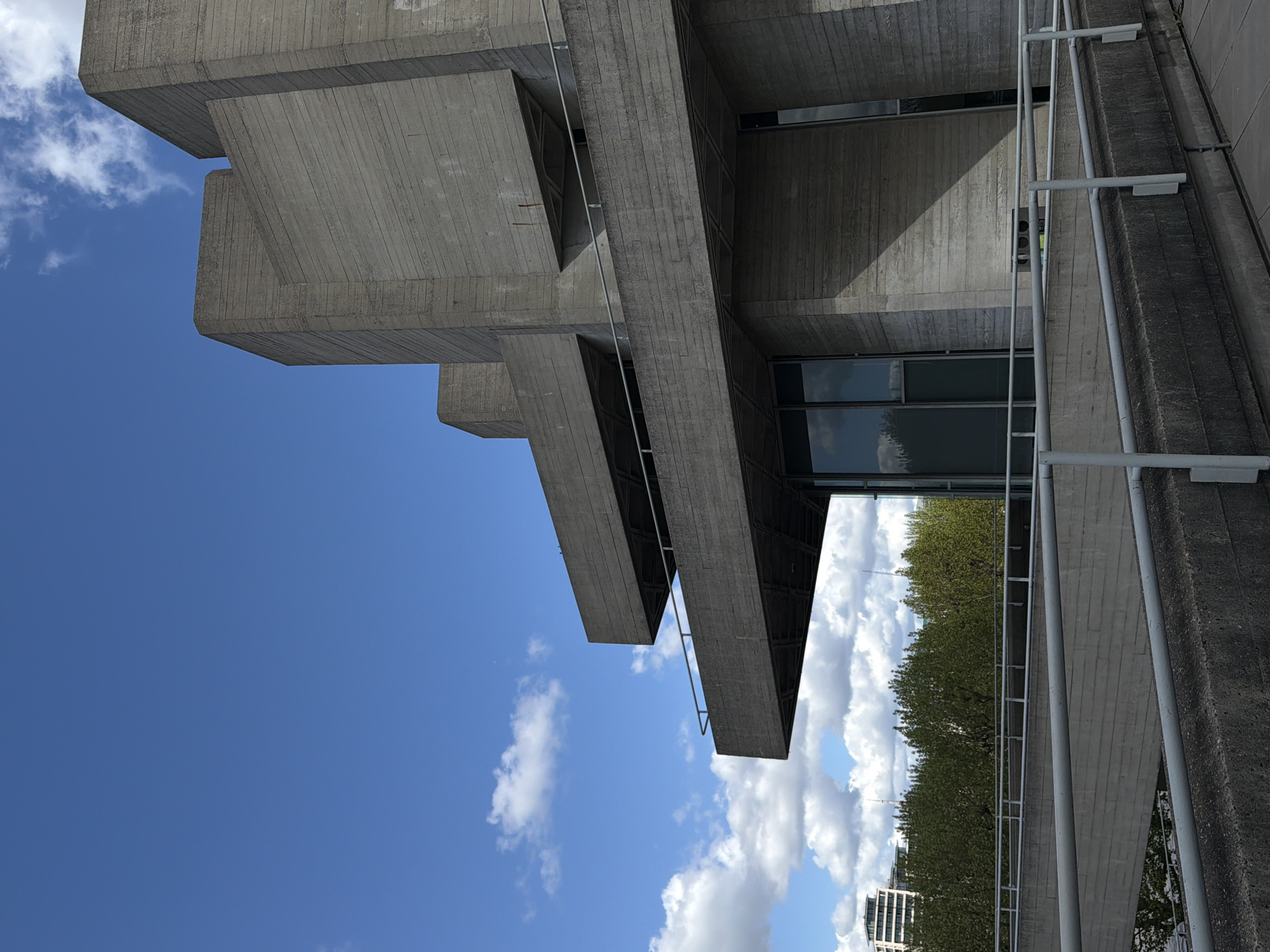 The image is a low-angle shot of a Brutalist concrete building against a blue sky with scattered white clouds. The building features cantilevered sections that create a layered effect, with visible horizontal striations suggesting concrete formwork. A metal railing runs along the side of the building, and a line of green trees is visible in the background. The building's windows reflect the sky and trees. The scene is located in London, United Kingdom, and the building is the Barbican Centre.