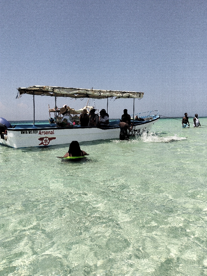 An image captured in Wasini, Kenya, depicts a white motorboat with blue trim anchored in shallow, clear, light blue-green ocean water under a bright, clear sky. The boat, labeled "WWFD/MS/050 Arsenal" and featuring the Arsenal FC cannon logo on its hull, has a temporary canopy structure providing shade.
Onboard the boat, at least five individuals are seated or standing. One person wears a wide-brimmed hat. An individual in dark clothing stands in the water near the boat's starboard stern, actively generating a splash. In the immediate foreground, a person with dark hair is floating face-down on a bright yellow-green flotation device. Further in the background, to the right of the boat, two male individuals stand waist-deep in the water; one wears white shorts, and the other wears dark shorts. The overall scene suggests recreational activity on a clear day.