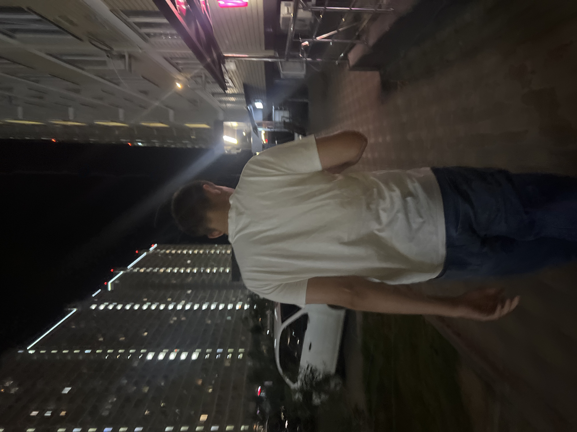 A person wearing a white t-shirt and blue jeans walks away from the camera on a paved sidewalk at night. To the left of the sidewalk are green bushes and a white car parked. Behind the bushes, a tall, illuminated apartment building with many windows is visible. To the right of the sidewalk is the side of another building, which appears to be a commercial establishment with an awning and neon signage. The overall scene depicts a residential area in the city of Krasnodar, Russia, at night.