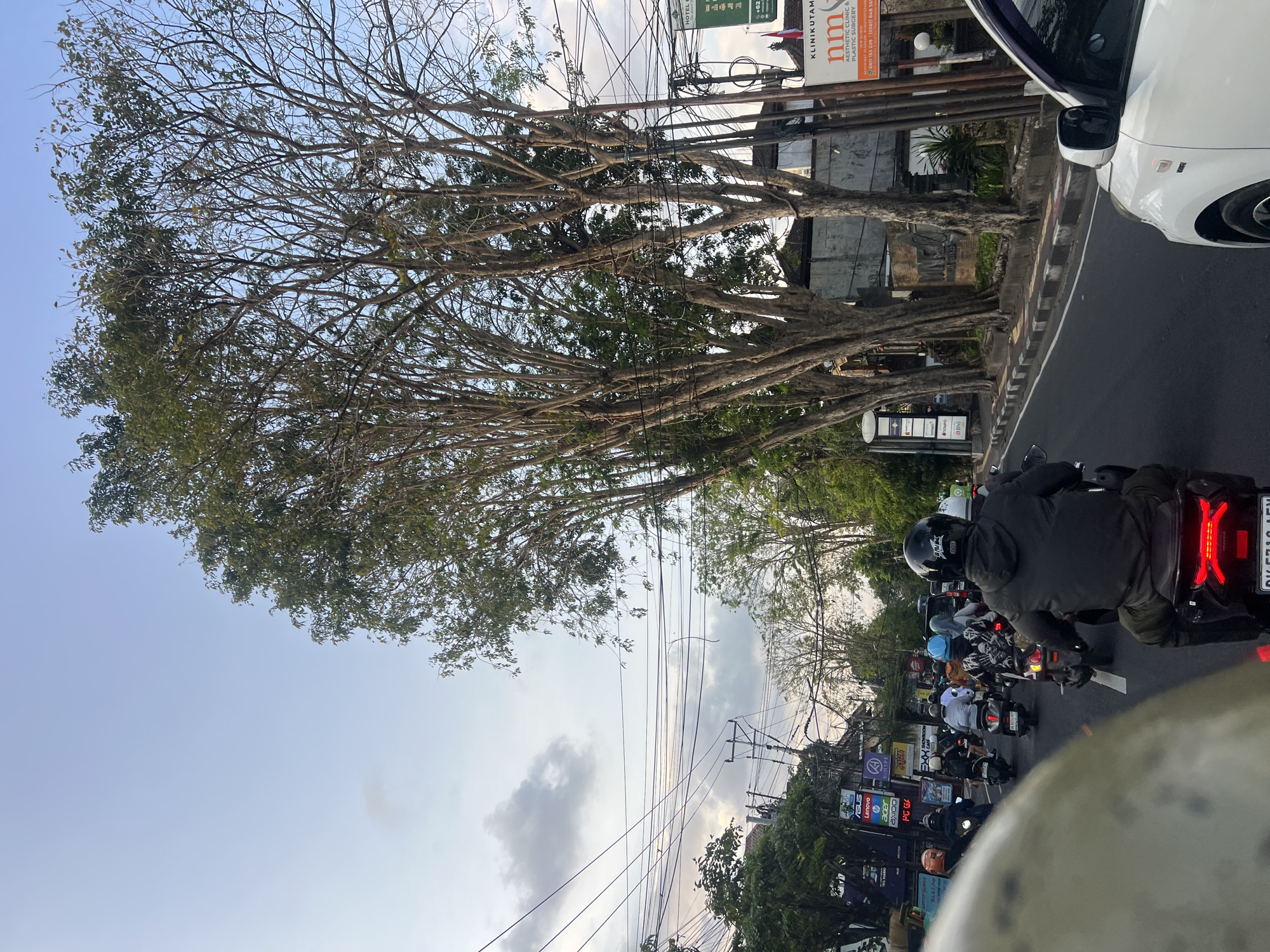 The image depicts a street scene in Denpasar, Indonesia, possibly during the late afternoon or early evening, given the lighting conditions and sky color. The viewpoint is from the perspective of someone on a motorcycle, looking forward at traffic.
In the foreground, the rear of a black motorcycle is visible, with its red taillight illuminated. The rider is wearing a black jacket and a black helmet with "DOT" marking. A portion of a white car is seen on the right side of the image, in the adjacent lane.
The road is lined with trees, some of which are tall and bare, while others have foliage. Power lines are strung across the street, with multiple utility poles visible. Along the side of the road, there are buildings and signs. One prominent sign reads "KLINIKUTAMA NM AESTHETIC CLINIC & PLASTIC SURGERY" with a phone number. Other smaller signs, some for brands like Asus and Lenovo, are also present on buildings further down the street.
A convoy of motorcycles is in motion on the road, indicating moderate traffic. Riders are wearing helmets of various colors, including black, white, and blue. Some are carrying passengers. The overall impression is of a busy urban environment with ongoing vehicular activity. The sky is partly cloudy with a light blue hue.
