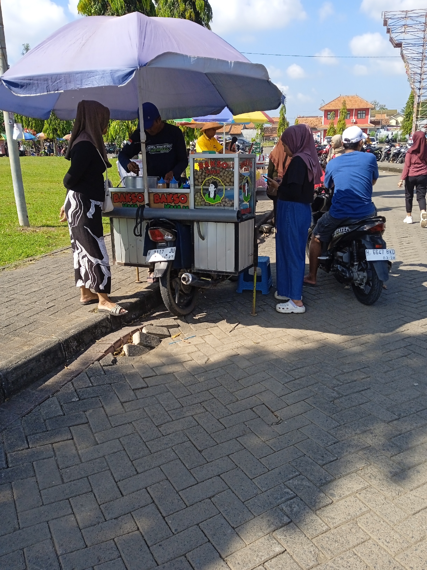 The image shows a street scene in Kebondalem, Indonesia. A food vendor operates a "Bakso" cart under a large light purple umbrella. Two customers stand on the left, while another customer is seated on a motorcycle on the right. The vendor is serving the customer while standing behind the cart. The street is paved with dark grey bricks. Behind the scene, there is a grassy area, buildings, and other motorcycles parked. The sky is blue with some clouds.