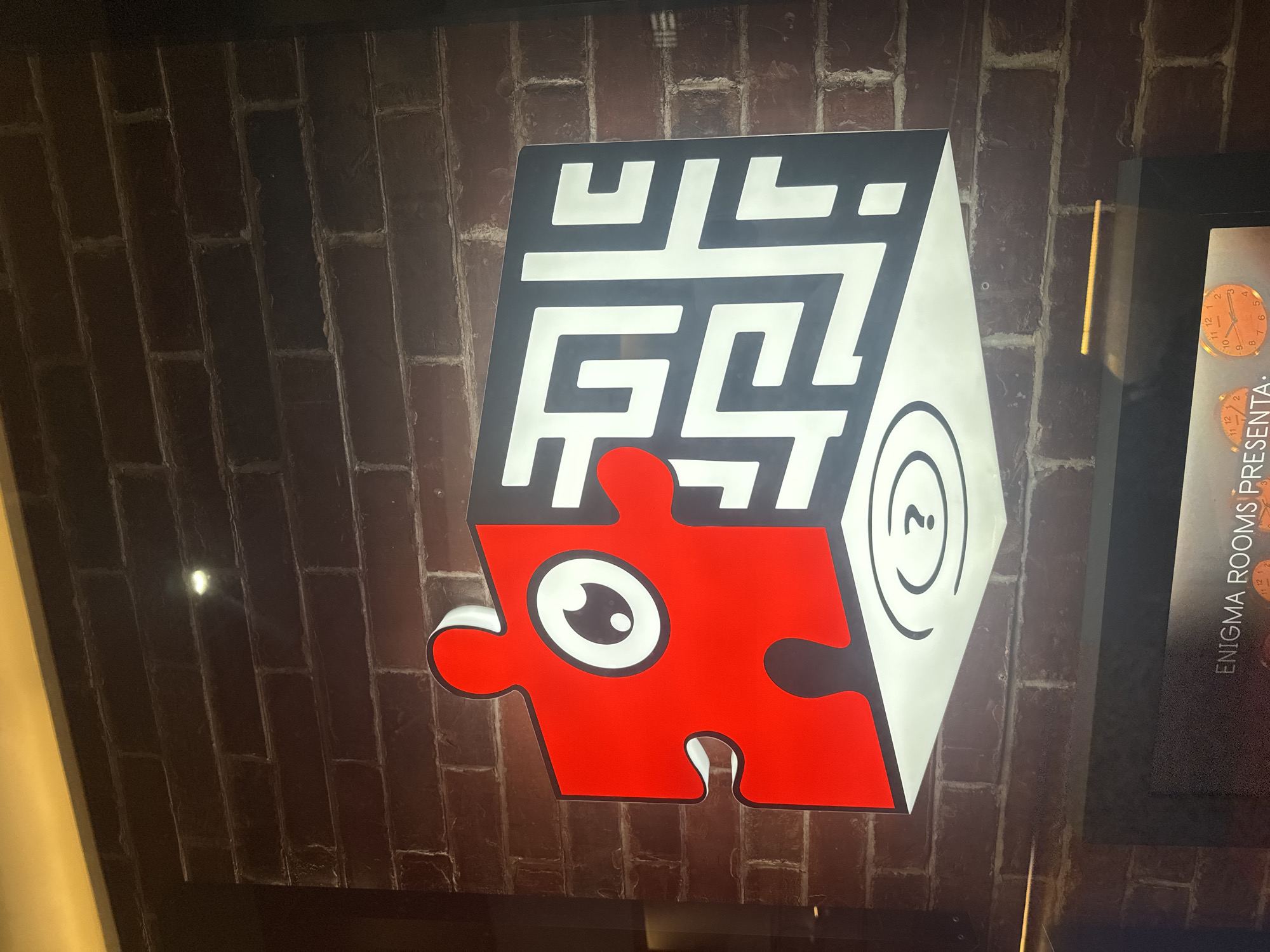A brightly lit sign is displayed on a brick wall. The sign features a gray cube with a white maze design on its top face. A red jigsaw puzzle piece with a white cartoon eye sits atop the cube. The side of the cube displays white concentric circles with a question mark in the center. To the right of the cube sign, a partial view of another sign is visible with the text "ENIGMA ROOMS PRESENTA:". The scene is illuminated, giving a clear view of the signs and the brick wall. Based on the text "ENIGMA ROOMS," this image likely depicts a promotional display for an escape room company, however, without more specific information, location cannot be precisely provided within Puebla, Mexico.