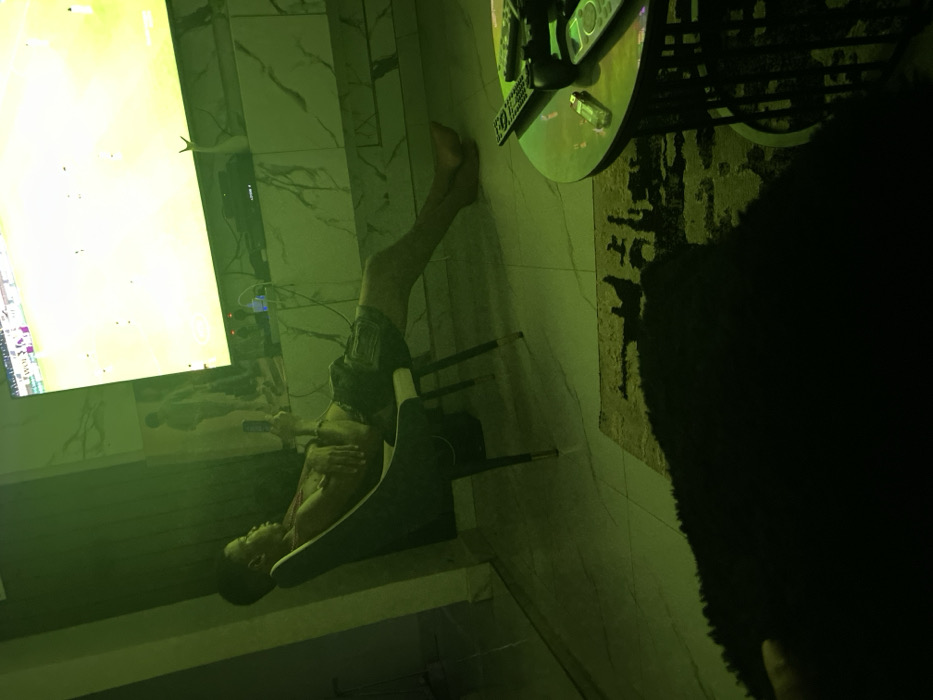 A shirtless person reclines in a black chair with their legs extended upward toward a large, bright television screen. The room is dimly lit with a green hue. A glass-topped table cluttered with remote controls sits nearby on a rug. The individual holds what appears to be a phone. The wall behind the TV has a marble pattern. A dark, obscured object fills the right side of the frame.