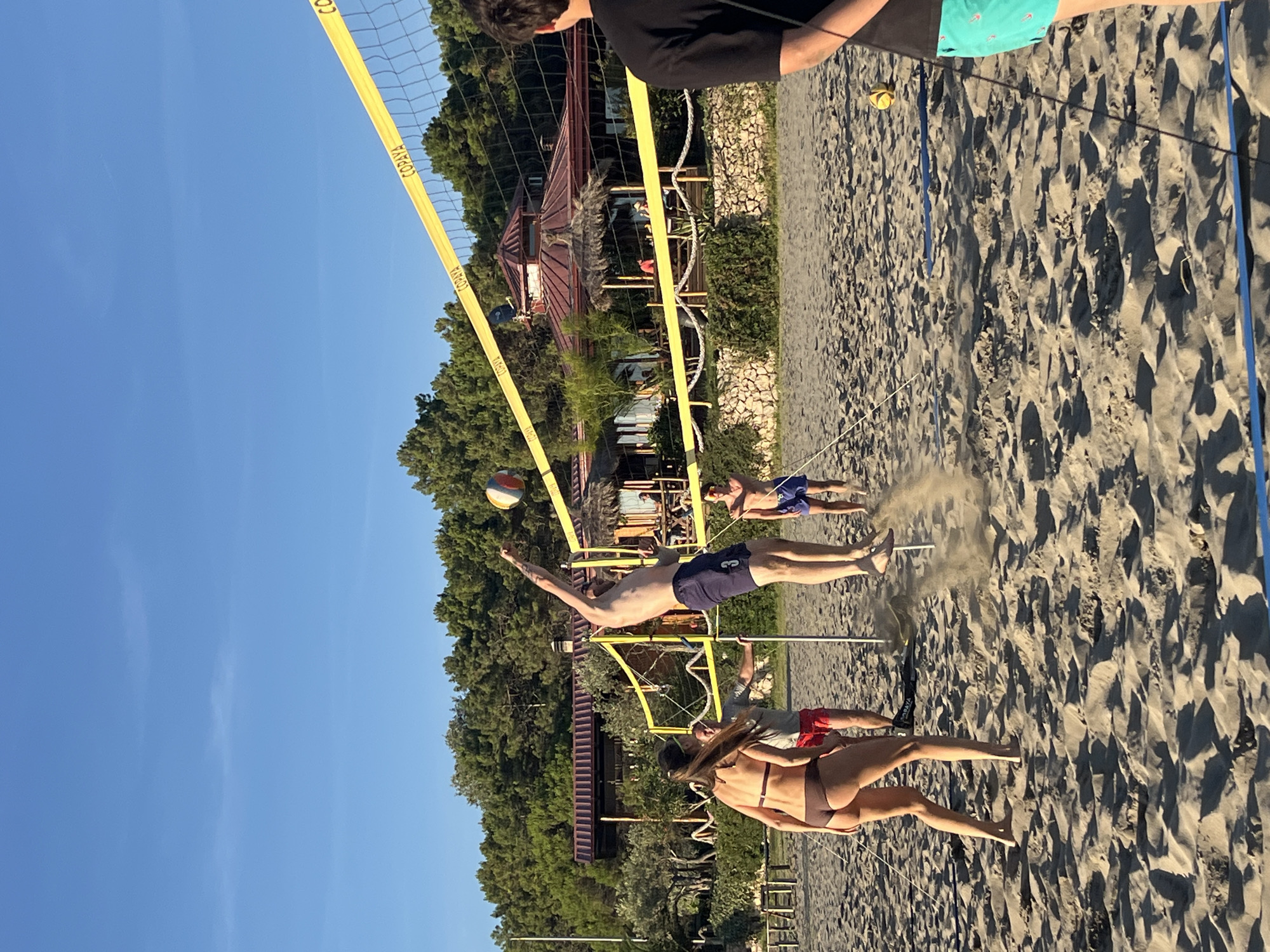 In Pistula, Montenegro, on a sandy beach, four individuals are playing beach volleyball. A man in blue shorts is mid-air, attempting to spike the ball over the net. A woman in a brown bikini is facing away from the camera, positioned on the left side of the court. Two other men are also on the court, one near the net and another further back. The game is taking place on a sunny day with a clear blue sky. Behind the volleyball court, there is a building with a reddish-brown roof and a thatched canopy, surrounded by green trees. A volleyball net with yellow tape runs across the middle of the court. The sand is textured with footprints and shadows from the sunlight.