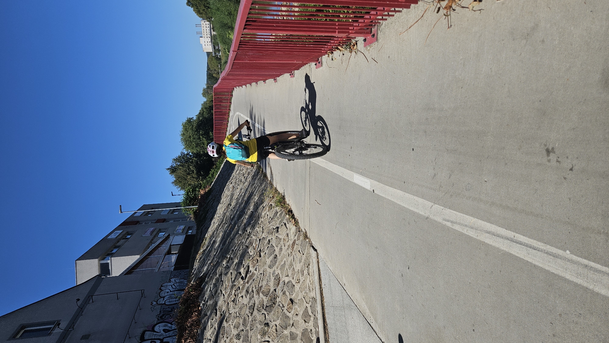 A person wearing a yellow shirt and a helmet with a blue backpack is riding a bicycle on a paved path next to a red railing. The path is marked with a white line. To the left of the path is a stone embankment, and beyond that, a grey building with graffiti. The background shows a clear blue sky, trees, and more buildings in the distance. This scene appears to be along the Torysa river embankment in Prešov, Slovakia, a popular location for cycling and walking.