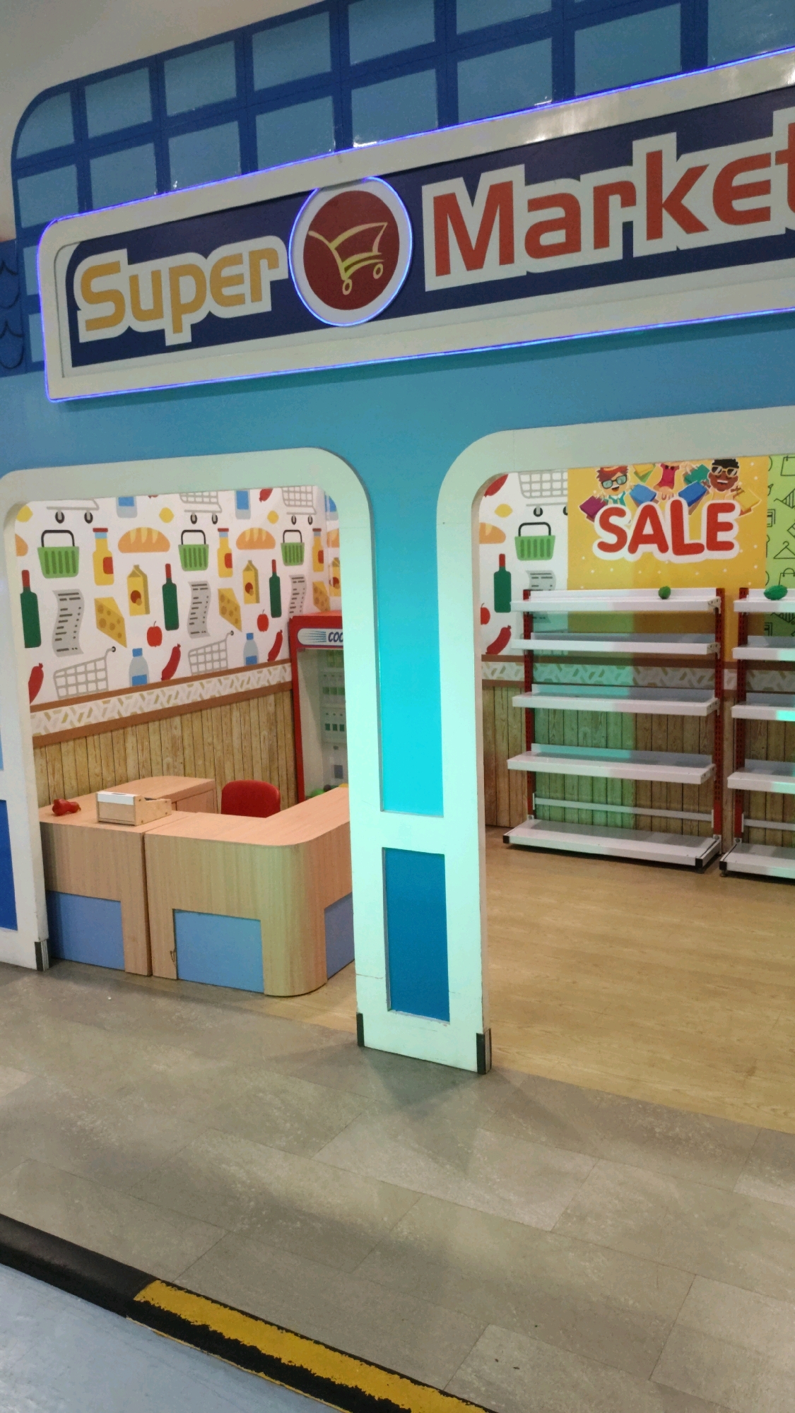 The image shows the interior of a brightly colored, stylized supermarket play area. A prominent sign at the top reads "Super Market" with a shopping cart icon. To the right, a banner announces "SALE" featuring cartoon children holding shopping bags. The play area includes shelves that appear to be stocked with toy products, and a counter area with a red stool. The walls are decorated with playful patterns of food items like bread, apples, and chili peppers, as well as shopping carts and baskets. The flooring transitions from a tiled area in the foreground to a wooden floor in the background. The overall environment is designed for children's play, resembling a miniature supermarket. There are no people visible in the image. This scene is located in a children's play area, possibly at an indoor playground or educational facility, but its specific location within the city of Bandung City, Indonesia cannot be determined from the image alone.