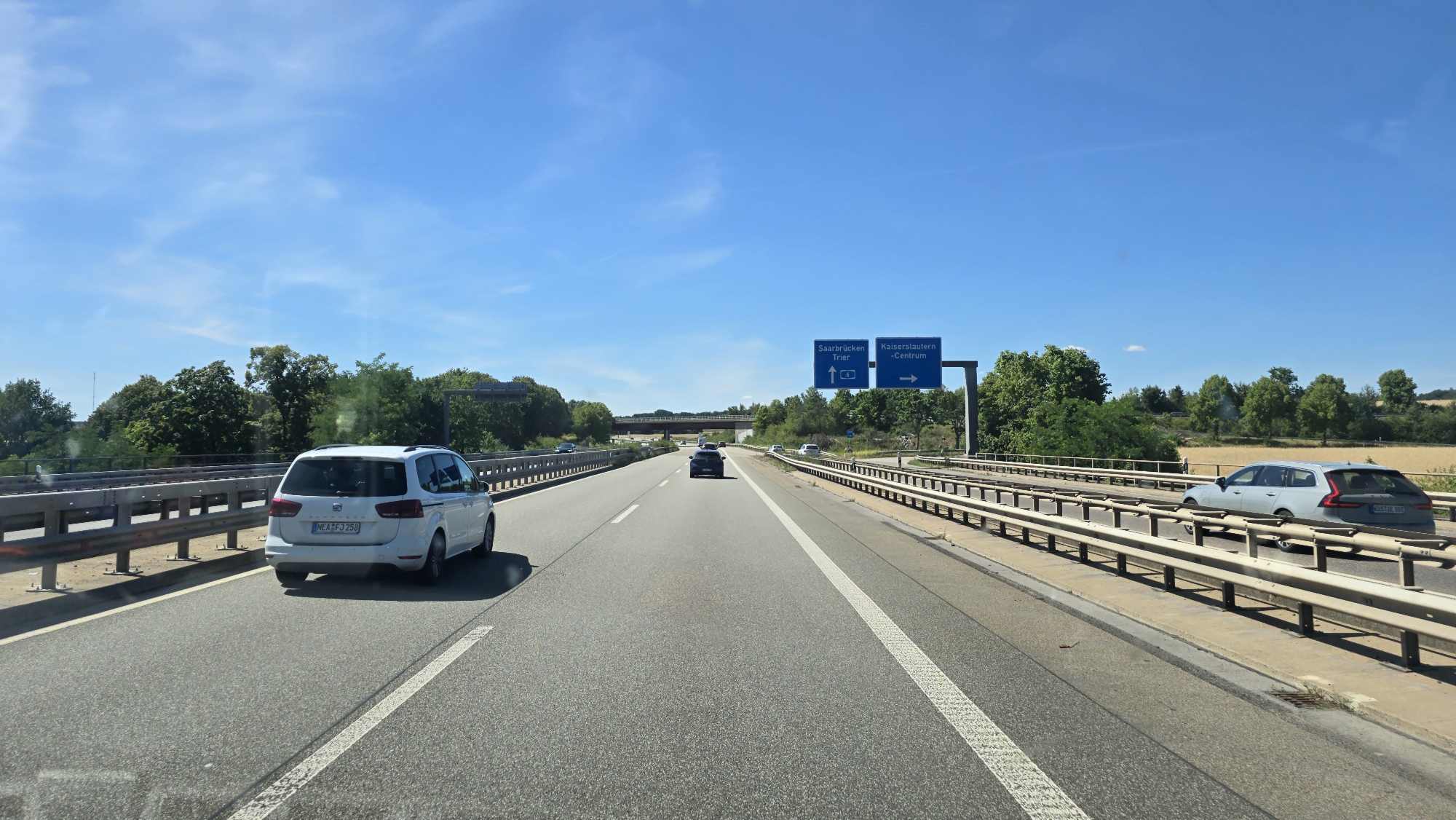 The image depicts a sunny day on a German autobahn. In the foreground, a white minivan with a license plate ending in "258" is driving on the right lane of the highway. In the middle ground, a dark-colored car is in the left lane. Further ahead, a blue road sign indicates directions to "Saarbrücken Trier" with an upward arrow and the number "6," and to "Kaiserslautern - Centrum" with a rightward arrow. The autobahn is flanked by metal guardrails and trees. In the far right, a light gray station wagon with a license plate ending in "88E" is visible. The sky is clear blue with a few wispy clouds. The surrounding environment appears to be rural, with fields and trees visible beyond the autobahn.