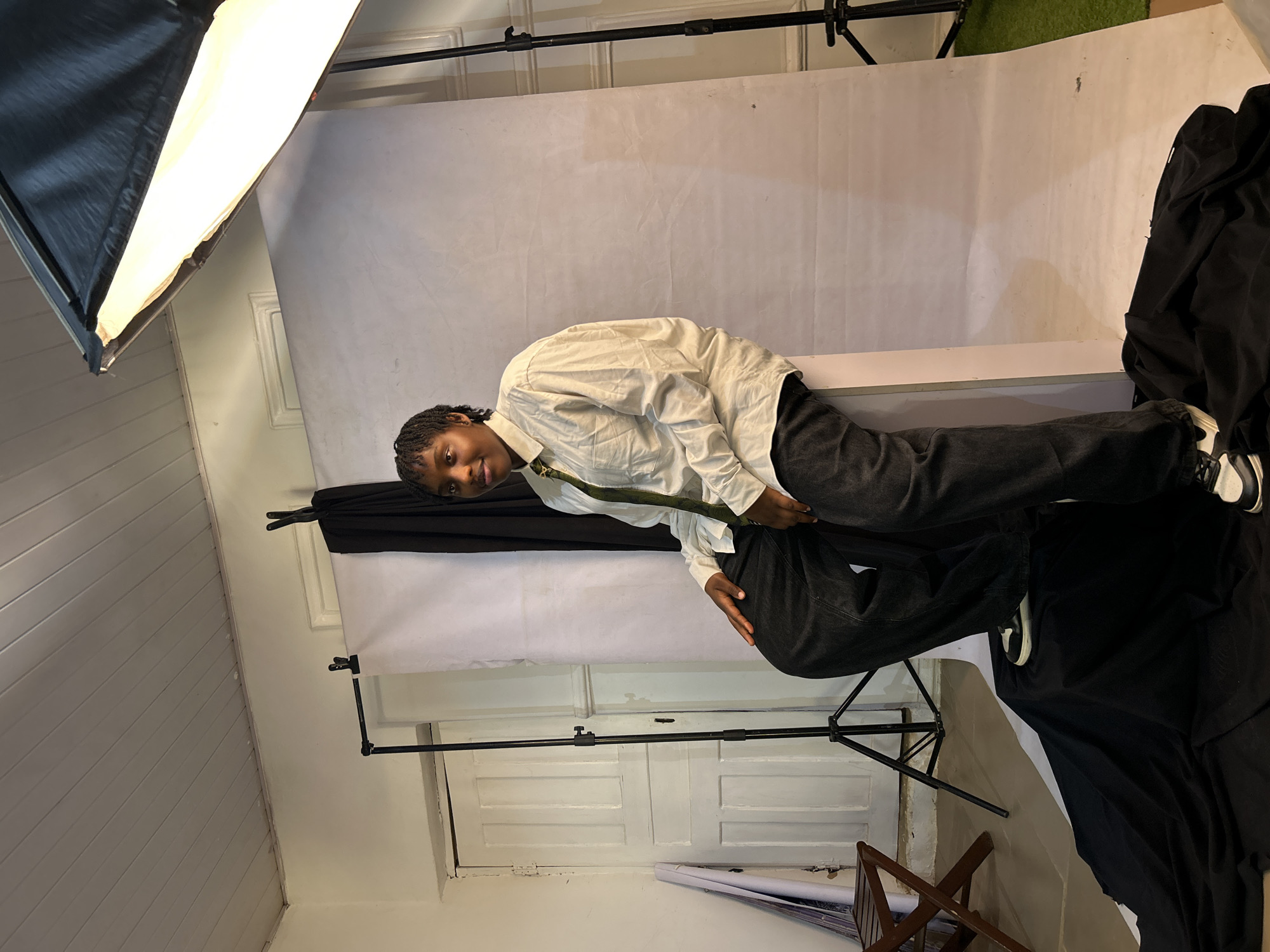 The image depicts a person with dark skin, dark braided hair, wearing a light-colored button-down shirt, dark pants, and sneakers, posing in a studio environment. The studio features a white backdrop partially covered with black fabric, lighting equipment on stands, and a white wall. A wooden folding chair is visible in the foreground. The location is most likely in an indoor studio space and there is no information available in the image to determine whether or not it is located in Jos, Nigeria.