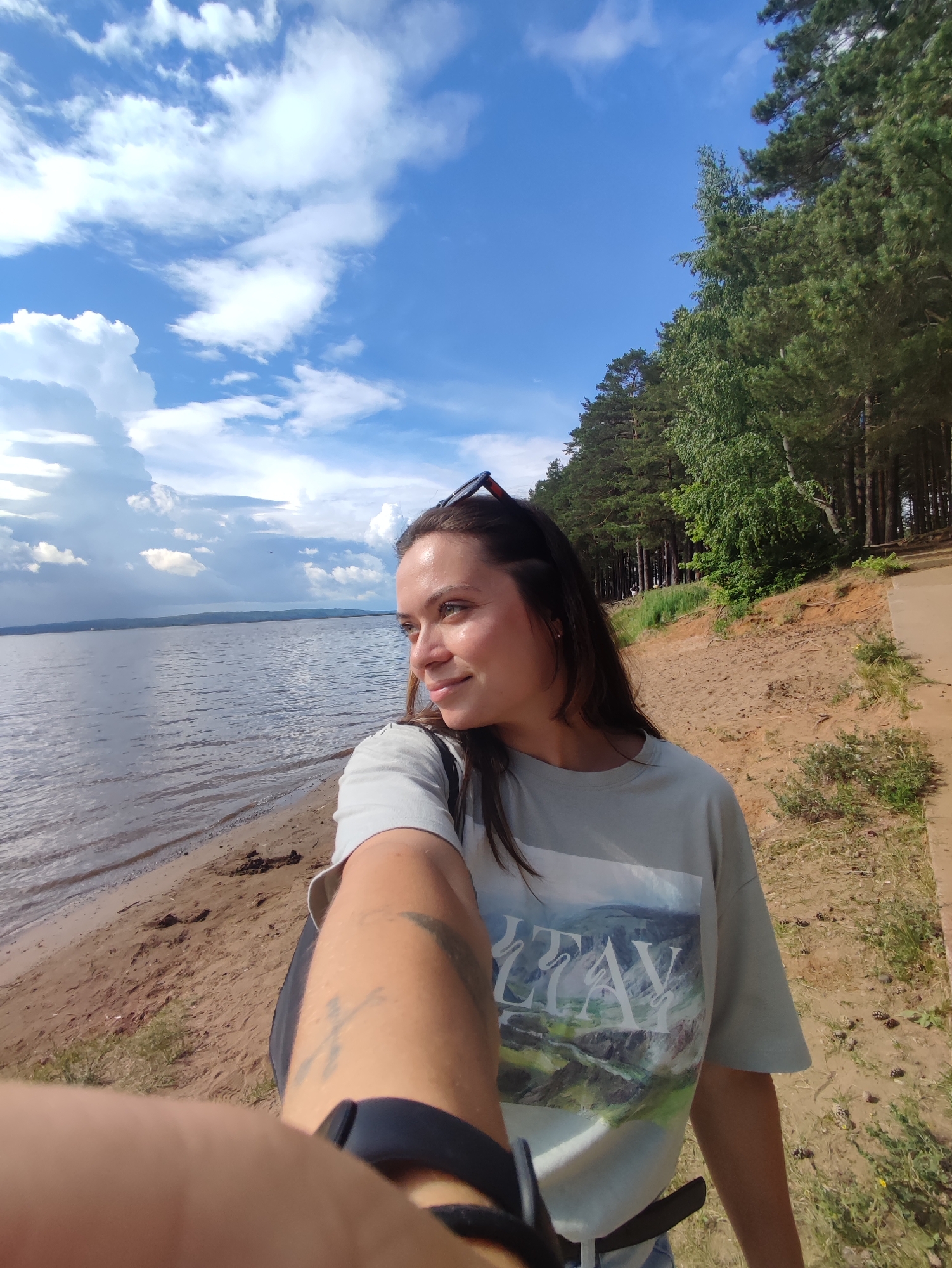 A young woman is captured in a selfie taken outdoors. She is wearing a light-colored t-shirt with a scenic landscape print. Sunglasses are perched on top of her head. The woman stands on a sandy beach beside a body of water, which appears to be the Kama River. Behind her, a dense forest of pine trees lines a sandy embankment. The sky is bright blue with scattered white clouds. The location is the bank of the Kama River in the city of Chaikovsky, Russia.